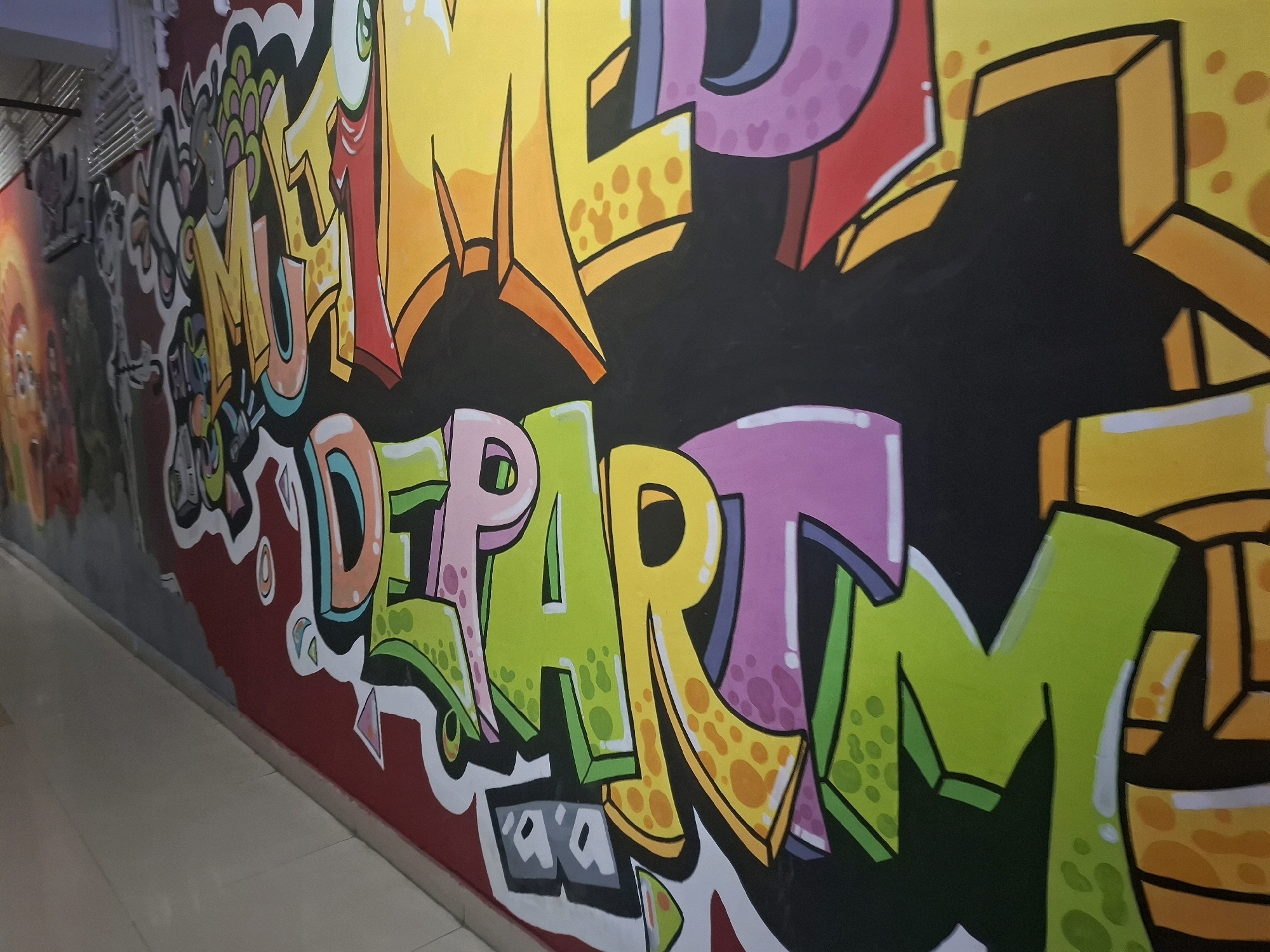 The image shows a vibrant graffiti mural on a wall. The mural features large, stylized letters in various bright colors including yellow, orange, pink, purple, and green. Some letters have a textured appearance with yellow dots. The background is predominantly black, contrasting with the colorful lettering. The graffiti is painted on an indoor wall, likely in a hallway or corridor, as suggested by the tiled floor and a railing or pipe running along the top left of the image. There are no people visible in the frame. The location is identified as Barasat, India, but this specific context is not visually evident from the image itself.