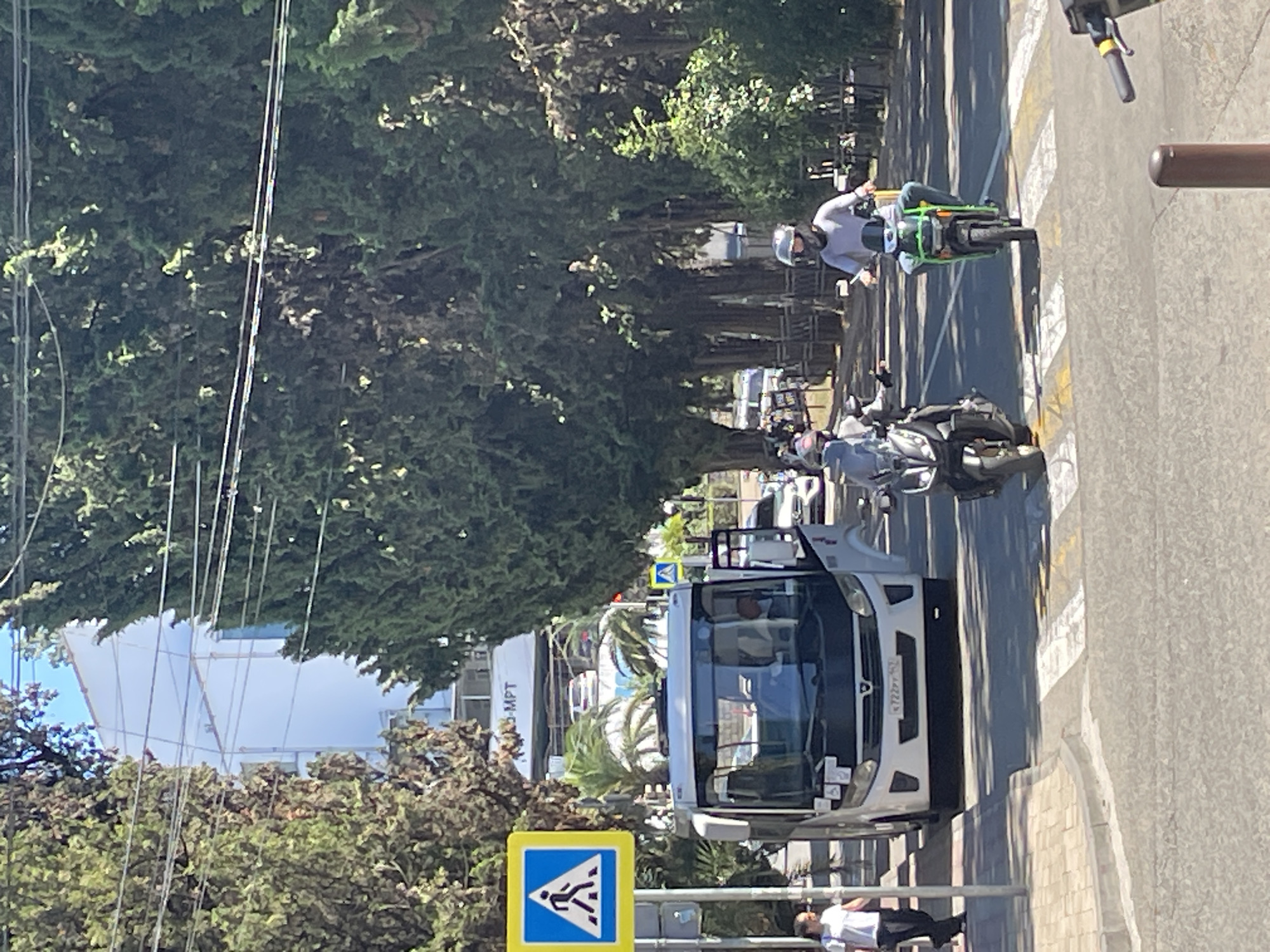 A street scene in Sochi, Russia, features a white bus stopped at a pedestrian crossing, with its door open. Two individuals on motorcycles are approaching or stopped at the crossing. One motorcycle rider, wearing a helmet and a grey shirt, is on a green scooter. The other motorcycle rider, also wearing a helmet, is on a darker-colored scooter. A pedestrian signal displaying a walking person is visible on the left. Tall trees line the right side of the street. In the background, a white building with scaffolding is visible, with a sign that appears to say "MPT". The road is paved, and there are painted lines for the pedestrian crossing and traffic lanes. The weather appears to be sunny.