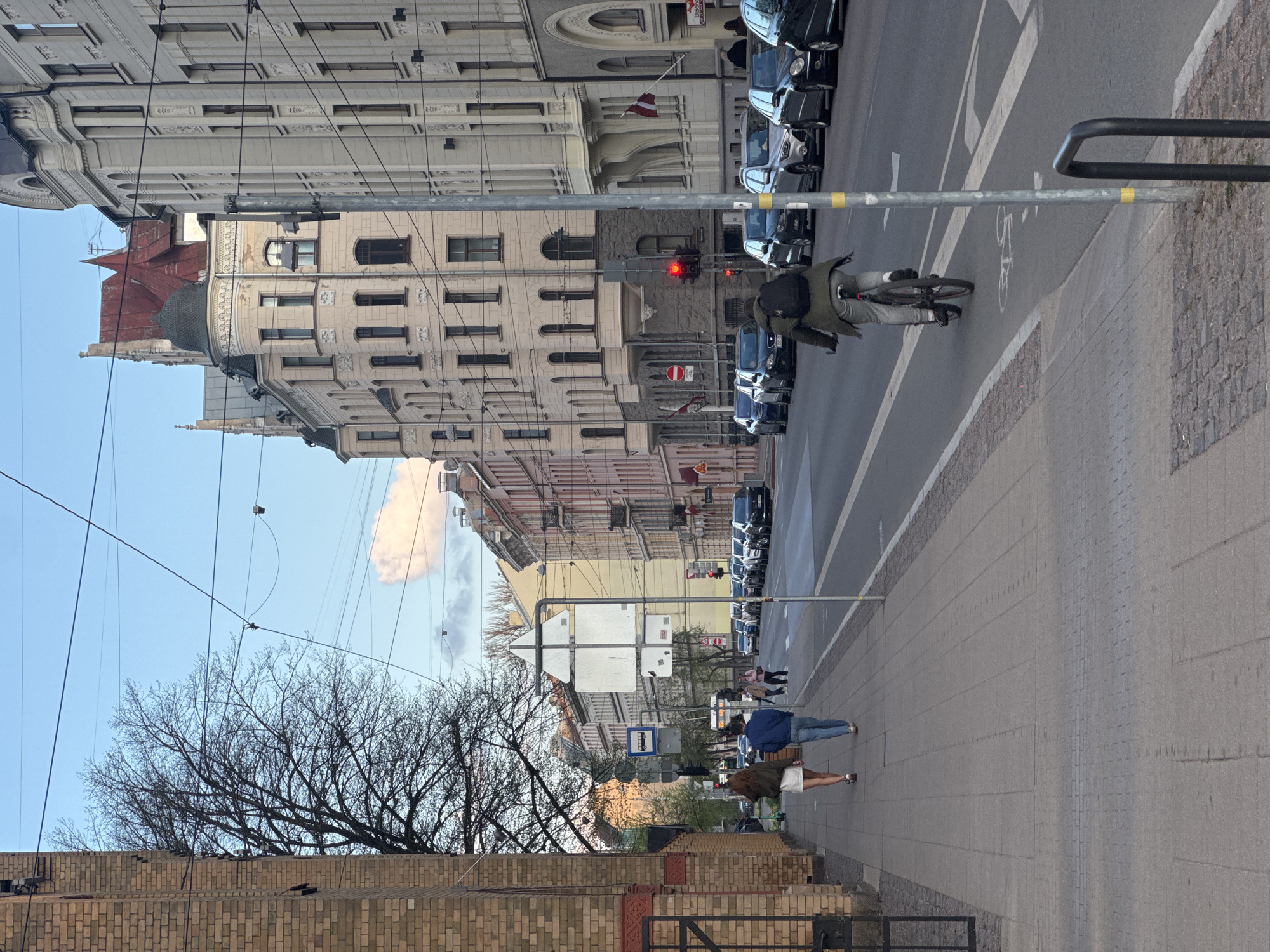 The image shows a street scene in Riga, Latvia. On the right, a wide gray-paved sidewalk stretches into the distance. Several people are walking on the sidewalk. A cyclist wearing a dark coat and light pants is riding on the street. Parked cars line the right side of the street. A red traffic light is visible. The architecture consists of multi-story buildings with light-colored facades, multiple windows, and some decorative elements. A Latvian flag can be seen on one of the buildings. Trees with bare branches are in the foreground on the left, with a brick wall beneath them. Overhead electrical wires are visible against a light blue sky with a cloud.