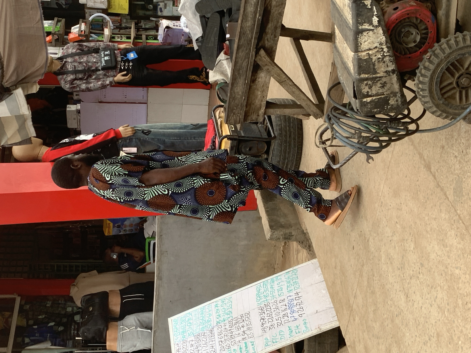This image captures a street scene in Shagamu, Nigeria, specifically in front of a shop with a red facade. In the foreground, a person wearing a vibrant, patterned African print outfit walks from left to right. The outfit consists of a long-sleeved tunic and matching trousers. The individual is wearing open-toed sandals. To the left of the person, a white board with handwritten text, likely lottery numbers or betting results, is propped up. Behind this board, mannequins are displayed, showcasing clothing items such as grey sweatpants and black shorts.
Further to the right, other mannequins are visible, one wearing a plaid shirt and a black chest bag with "Death Note" written on it, and another with a red and black long-sleeved shirt. In the middle ground, a portable generator sits on a tire, with a coiled hose nearby. A wooden table or stand, seemingly used for display, is also present. The background shows the interior of the shop, with shelves stocked with various items. The overall scene depicts a bustling commercial area with elements of retail, public display, and daily commerce.