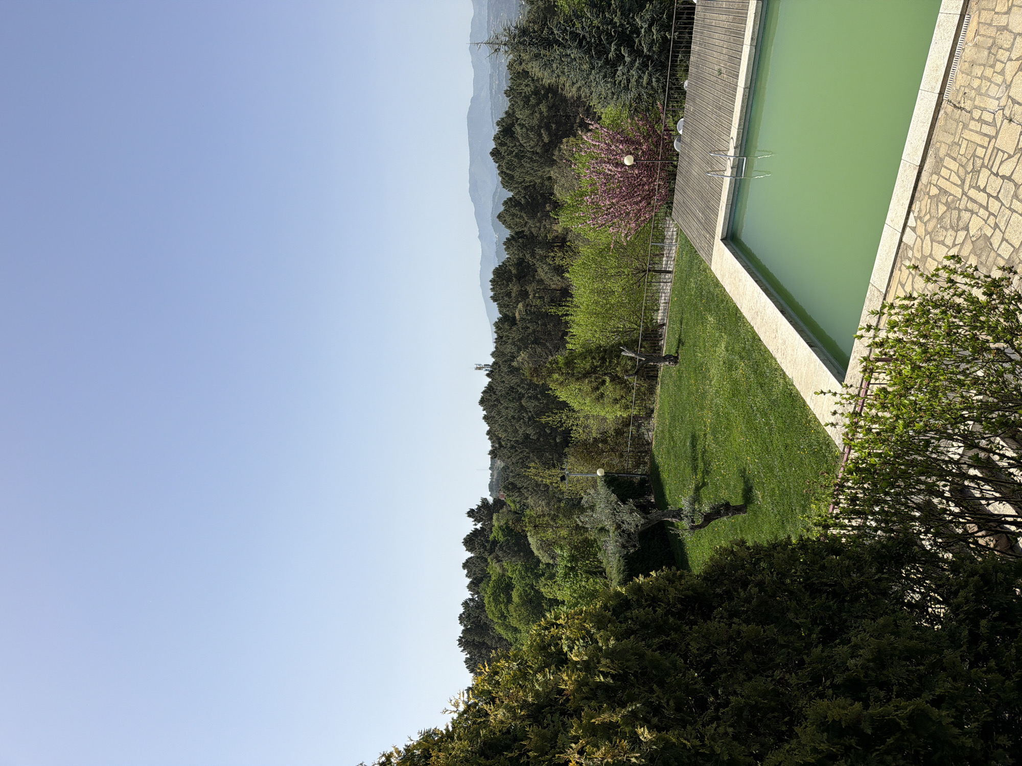 The image shows a landscape with a swimming pool, trees, and a distant mountain range under a clear blue sky. The swimming pool has green water, and a wooden deck surrounds part of it. There is a well-maintained lawn beside the pool. The trees are lush and green, with some flowering trees adding pops of color. In the distance, a mountain range is visible. The location is Vila Real, Portugal.