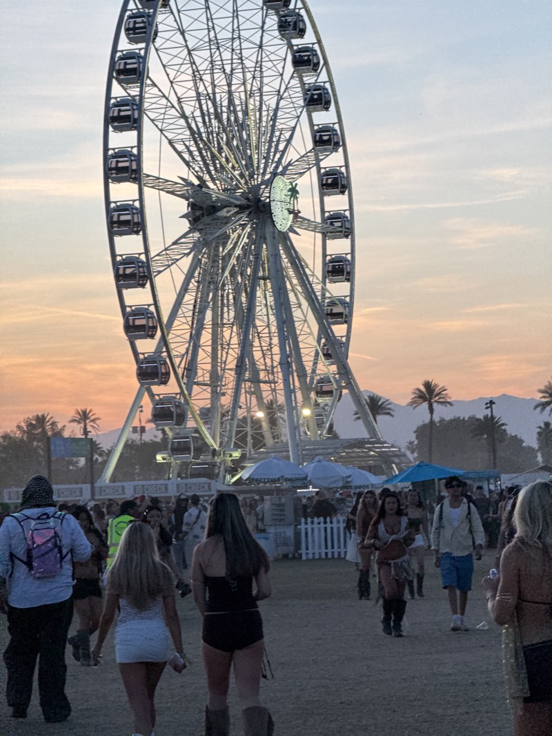 The image captures a large Ferris wheel as the focal point of an event. The Ferris wheel is towering and illuminated, with a design at its center, possibly a logo or symbol. Below it, a crowd of people strolls through the event space. Many of the people are dressed in casual summer attire and appear to be attendees of a music festival.
In the background, palm trees dot the horizon, and what appears to be a distant mountain range can be seen. The sky is light with warm hues suggesting it is either sunrise or sunset. The scene conveys the atmosphere of an outdoor festival or event, likely taking place at dusk or dawn in a warm climate. Based on the presence of palm trees and Ferris wheel this location is in the city of Indio in the United States.