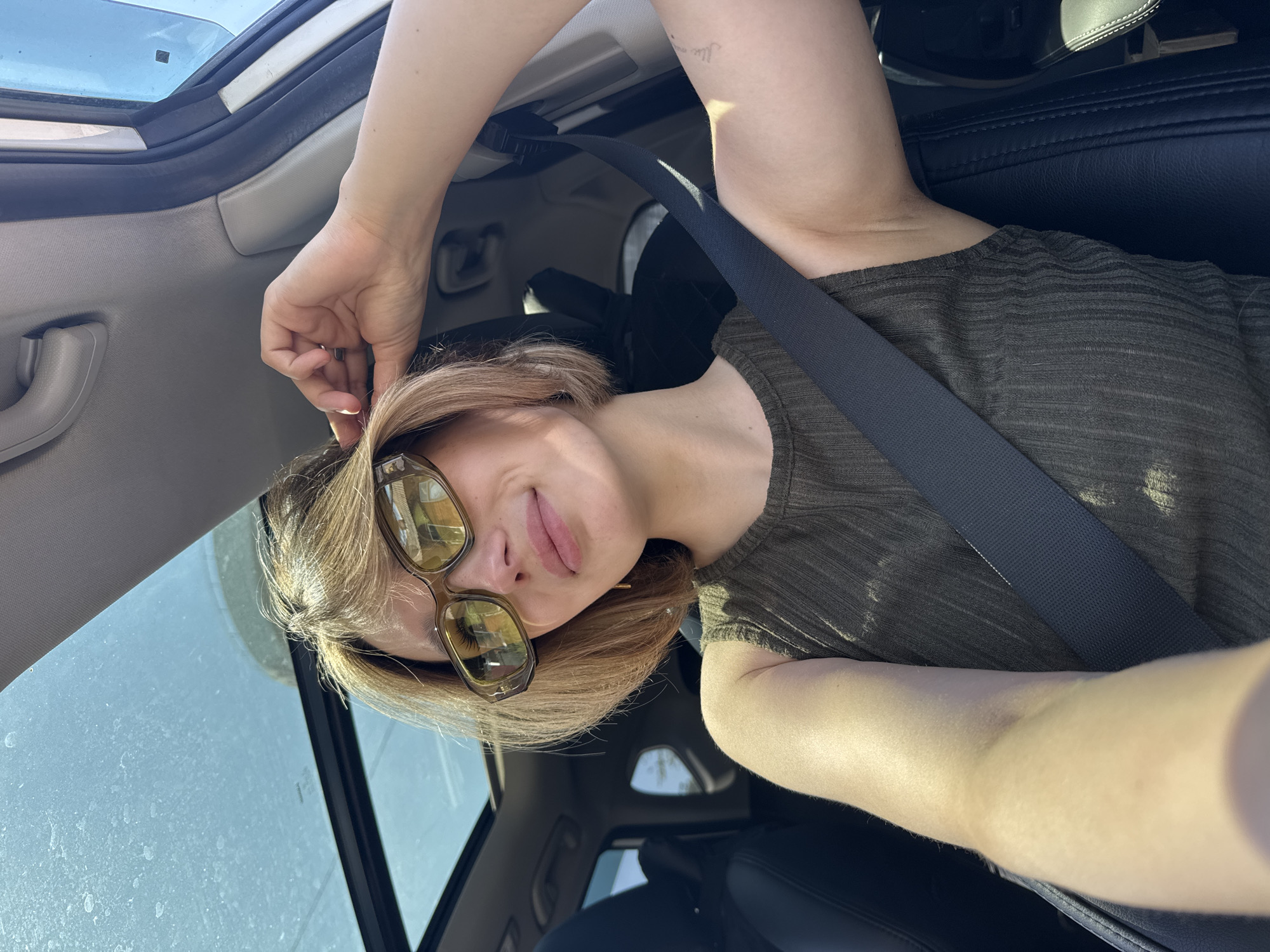 A woman with short blonde hair and sunglasses is sitting in the driver's seat of a car with a sunroof. She is wearing a dark green sleeveless shirt and a seatbelt. Her left hand is touching her hair, and her right arm is resting on the seatbelt. The car appears to be parked, and the scene is outdoors, illuminated by sunlight.