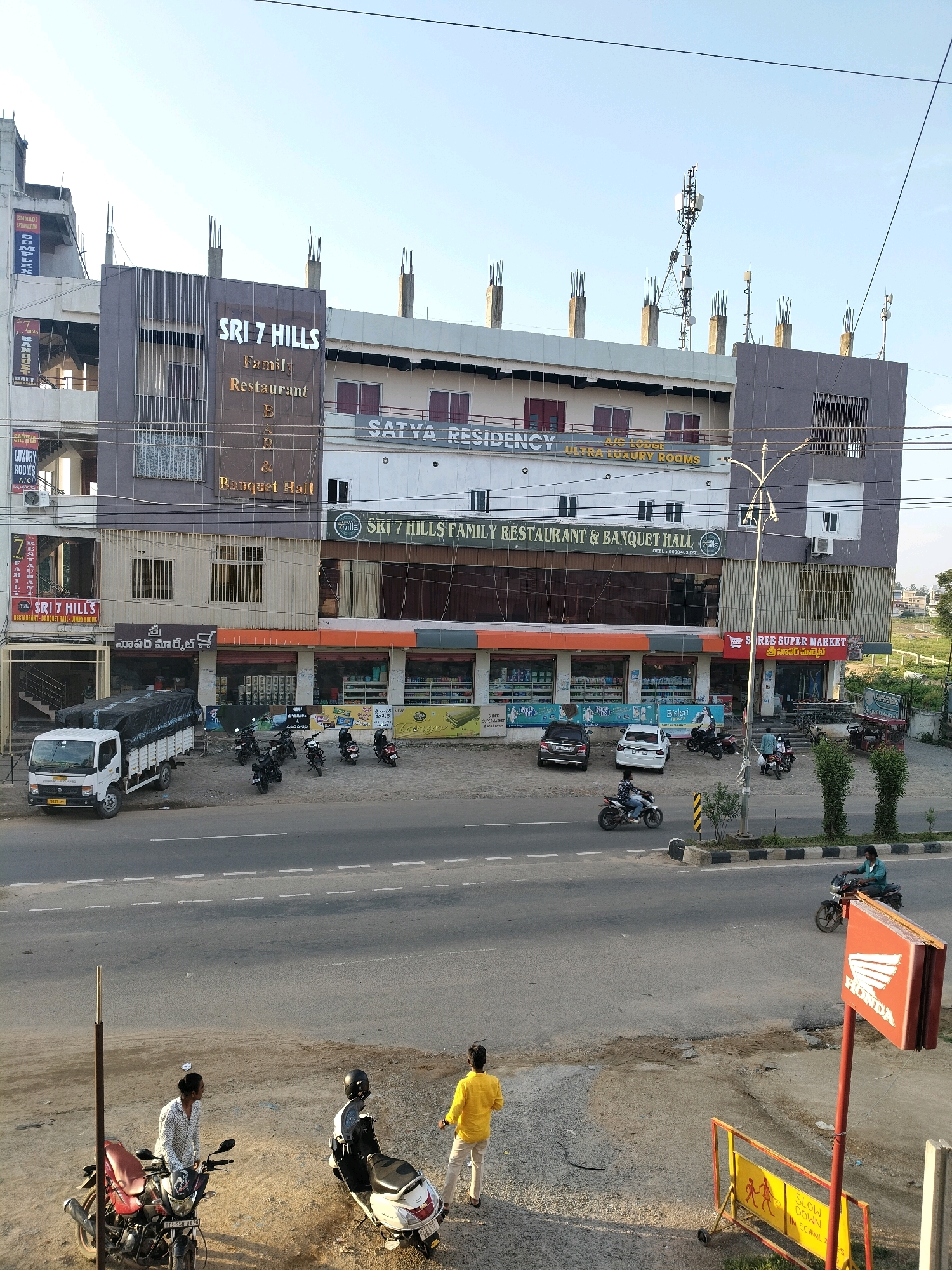 The image depicts a street scene in Sitarampur, India, with a multi-story building prominently featured. The building's facade displays signage for "SRI 7 HILLS FAMILY RESTAURANT & BANQUET HALL" and "SATYA RESIDENCY A/C LOUNGE ULTRA LUXURY ROOMS." Several businesses are visible at the ground level, including what appears to be a supermarket with products displayed through windows and another signage that reads "SHREE SUPER MARKET". A white delivery truck is parked in front of one of the establishments. The street in the foreground is paved and has white lane markings. Several motorcycles and scooters are parked along the roadside, and a few individuals are present on foot near the motorcycles. Some are standing on the sidewalk area, while others are in the process of riding or dismounting their vehicles. In the background, there is a telecommunications tower and other construction activities are evident on the top floors of some buildings. The sky is clear and the lighting suggests it is daytime.
