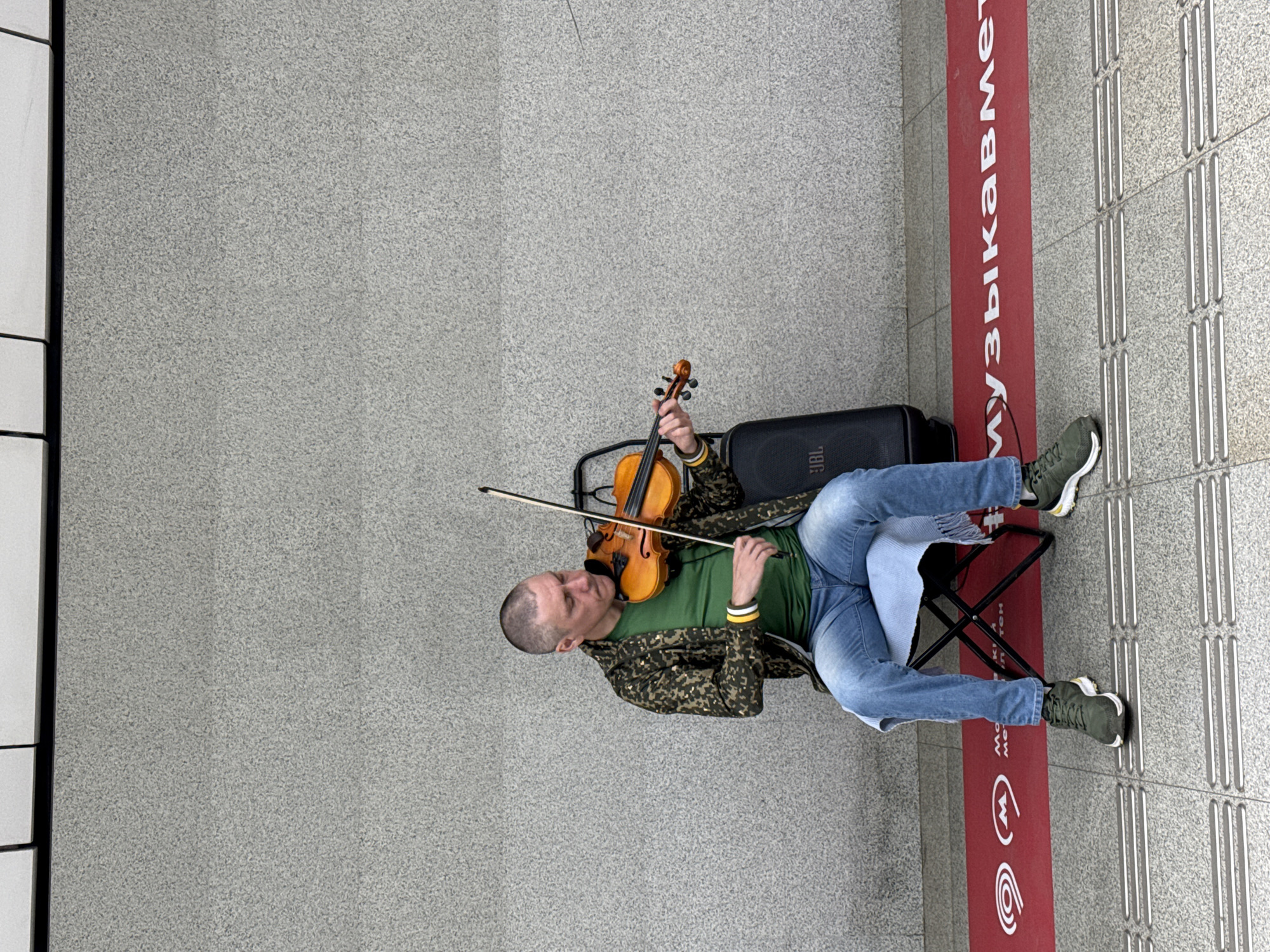A man is playing a violin while sitting on a folding stool in front of a grey, textured wall. He is wearing a green t-shirt, a camouflage jacket, blue jeans, and dark green sneakers. A black JBL speaker is positioned behind him. A red mat with white text and symbols is on the ground in front of him. The man is looking down at the violin as he plays. The scene appears to be in a public space, possibly a subway station, given the mat with text that appears to be in Russian.