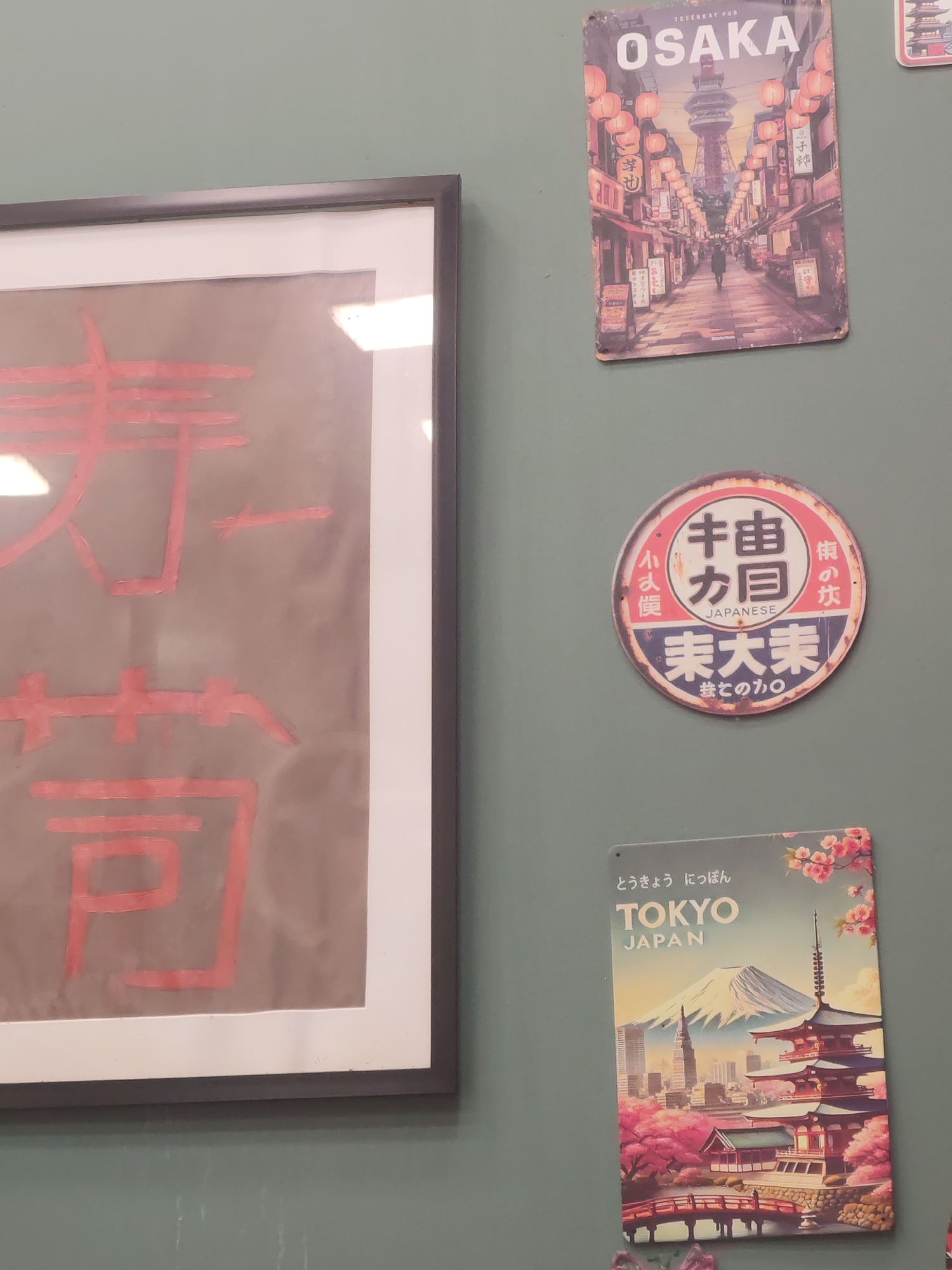 The image shows a wall with several decorative items. On the left, there is a framed piece of art with red calligraphy on a dark background. To the right of the artwork, there are three metal signs: an "OSAKA" sign depicting a street scene with lanterns and a tower, a circular sign with Japanese text and the word "JAPANESE" in English, and a "TOKYO JAPAN" sign featuring Mount Fuji, cherry blossoms, and a pagoda. The wall appears to be painted a muted green. There are no people visible in the image. The scene is not identifiable as being in the city of Baku, Azerbaijan.