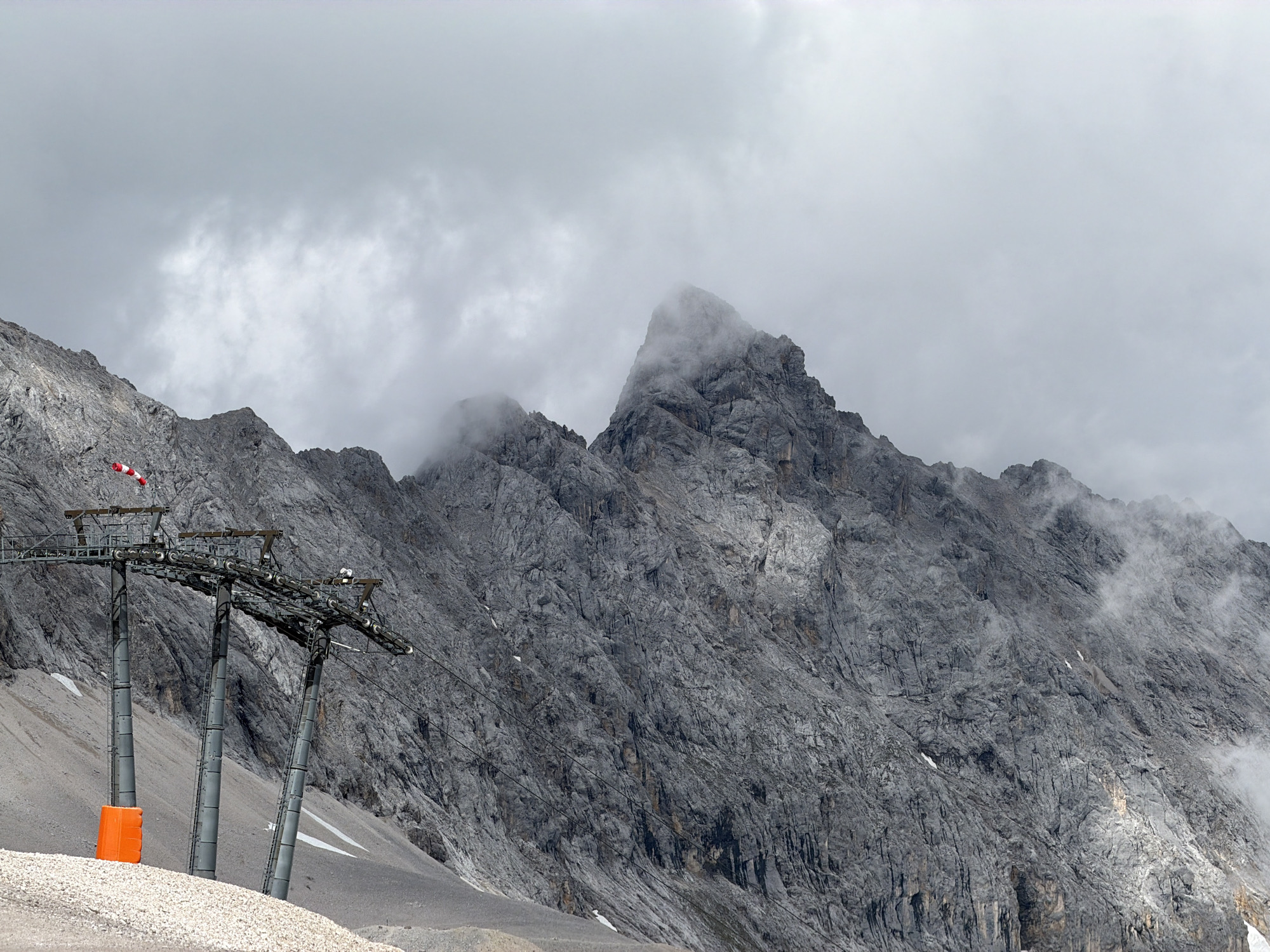 The image captures a dramatic mountain landscape near Garmisch-Partenkirchen, Germany, under a cloudy sky. In the foreground, a ski lift support tower stands tall, its metal structure a stark contrast against the rugged, grey rock face of the mountain behind it. A bright orange protective barrier is visible at the base of one of the tower's legs, and a windsock with red and white stripes flutters from the top of the structure. The mountain itself is massive and imposing, with deep fissures and varying textures carved by time and elements. Wisps of fog and clouds cling to the peaks, partially obscuring their summits and adding to the atmospheric and slightly ominous feel of the scene. The ground in the immediate foreground is covered in loose, light-colored gravel and small stones.
