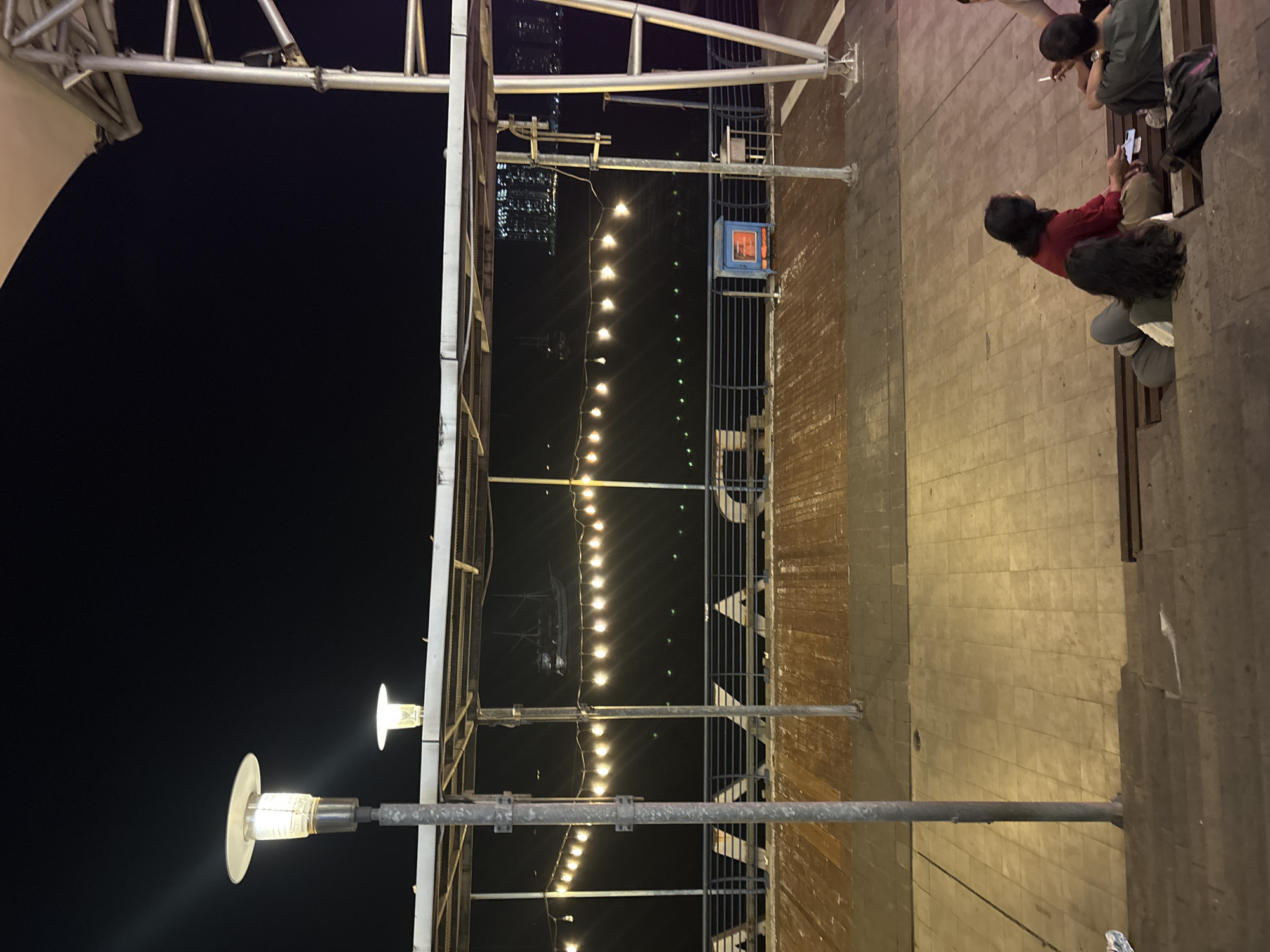 In North Jakarta, Indonesia, at night, a group of people are sitting on steps near a body of water. A wooden deck area is visible in the background, bordered by a railing and illuminated by string lights. A tall lamppost stands on the left side of the image, and another shorter lamp is positioned on a railing in the distance. The scene is lit by artificial lights from the lamps and string lights, casting a warm glow on the area. The dark sky suggests it is late evening or night.
