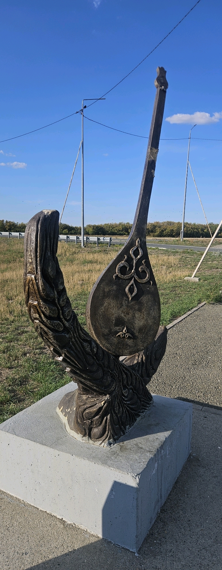 This is a medium-shot, eye-level, vertical, outdoor photograph of a sculpture of a dombra, a traditional Kazakh stringed instrument, situated on a concrete pedestal. The sculpture, made of a dark, weathered material, features intricate detailing on the body of the dombra, resembling swirling patterns. The base of the sculpture appears to be shaped like wings or flowing fabric. The sculpture is positioned in an open, grassy area with a paved path visible to the right. In the background, a road with a concrete barrier and distant trees under a clear blue sky are present. Power lines and light poles are also visible in the background. The location is specified as Қалбатау ауылдық округі, Kazakhstan. There are no people or animals visible in the frame. The lighting suggests it is daytime, likely morning or afternoon, with a strong shadow cast by the sculpture on the concrete base.