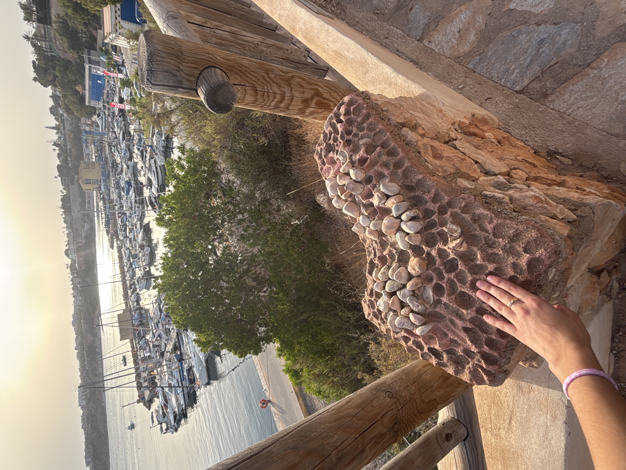 In Urbanización Cabo Roig, Spain, a hand with a ring and a pink bracelet rests on a textured, reddish-brown rock adorned with small, smooth stones. The rock, part of a low wall or structure, sits near a wooden fence. In the background, a marina is filled with numerous sailboats and yachts, with buildings and greenery lining the shore and a hazy, sunlit sky above.