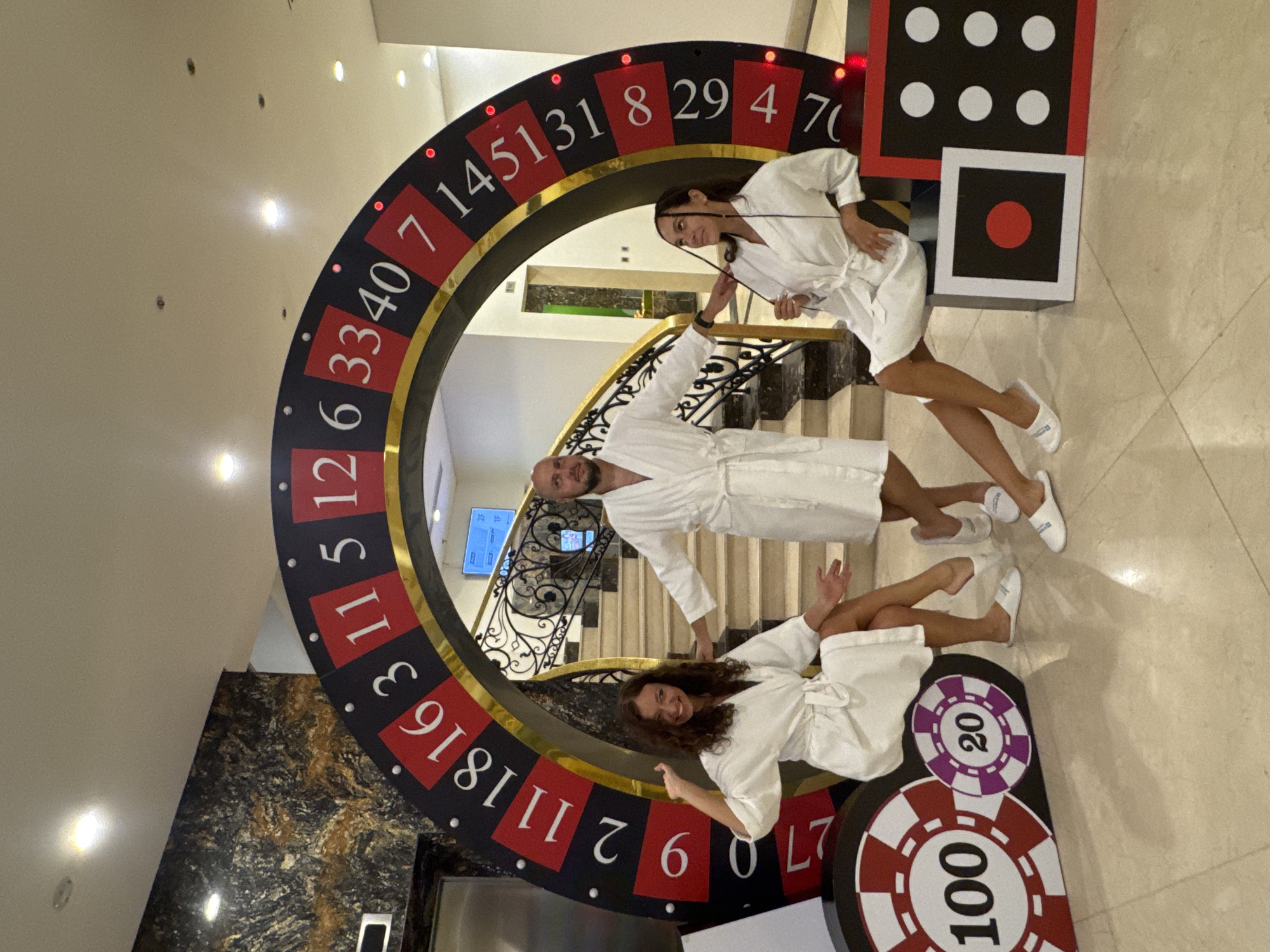 The image shows three people dressed in white bathrobes posing around a casino-themed setup, which includes a large roulette wheel, dice, and poker chips. The roulette wheel has numbers in red and black, ranging from 0 to 70, and is decorated with lights. The dice props are black with white dots and red outlines. The poker chips are large, round, and feature the numbers 100 and 20 in red and purple.
The man is standing in the middle, his bathrobe wrapped around him. One of the women is leaning on the roulette wheel at the bottom, and the other woman is standing at the top.
The scene appears to be indoors, possibly in a hotel or resort, given the elegant staircase with wrought-iron railings visible in the background. The setting also indicates a location in a city, and based on the image's context and available information, the city is Đà Nẵng, Vietnam.