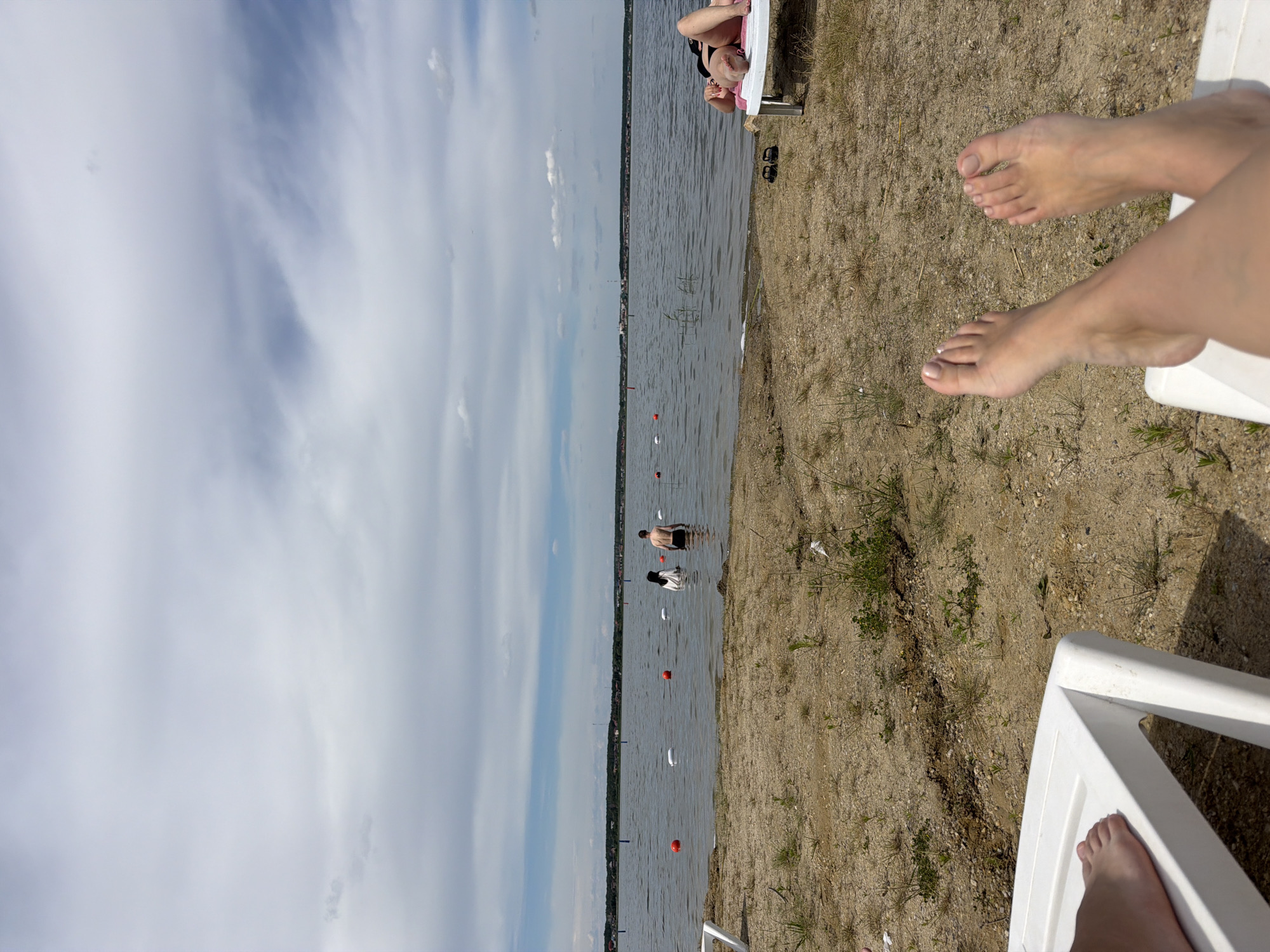 The image depicts a section of a sandy beach on a cloudy day, likely at the New Kremenkul' reservoir in the city of New Kremenkul', Russia. In the foreground, the legs and feet of a person are visible, resting on a white lounge chair. The sand is sparsely covered with dry grass and small plants. In the middle ground, a body of water, the reservoir, stretches across the frame. Several people are present in the water. Two individuals, a man and a woman, are standing partially submerged, with the woman wearing a headscarf. Further in the water, additional figures can be seen. Orange buoys are scattered across the water's surface, marking a swimming area. On the right side of the frame, another person is lying on a pink-cushioned lounge chair, also facing the water. The background shows a distant shoreline with buildings and trees under a sky filled with scattered clouds.