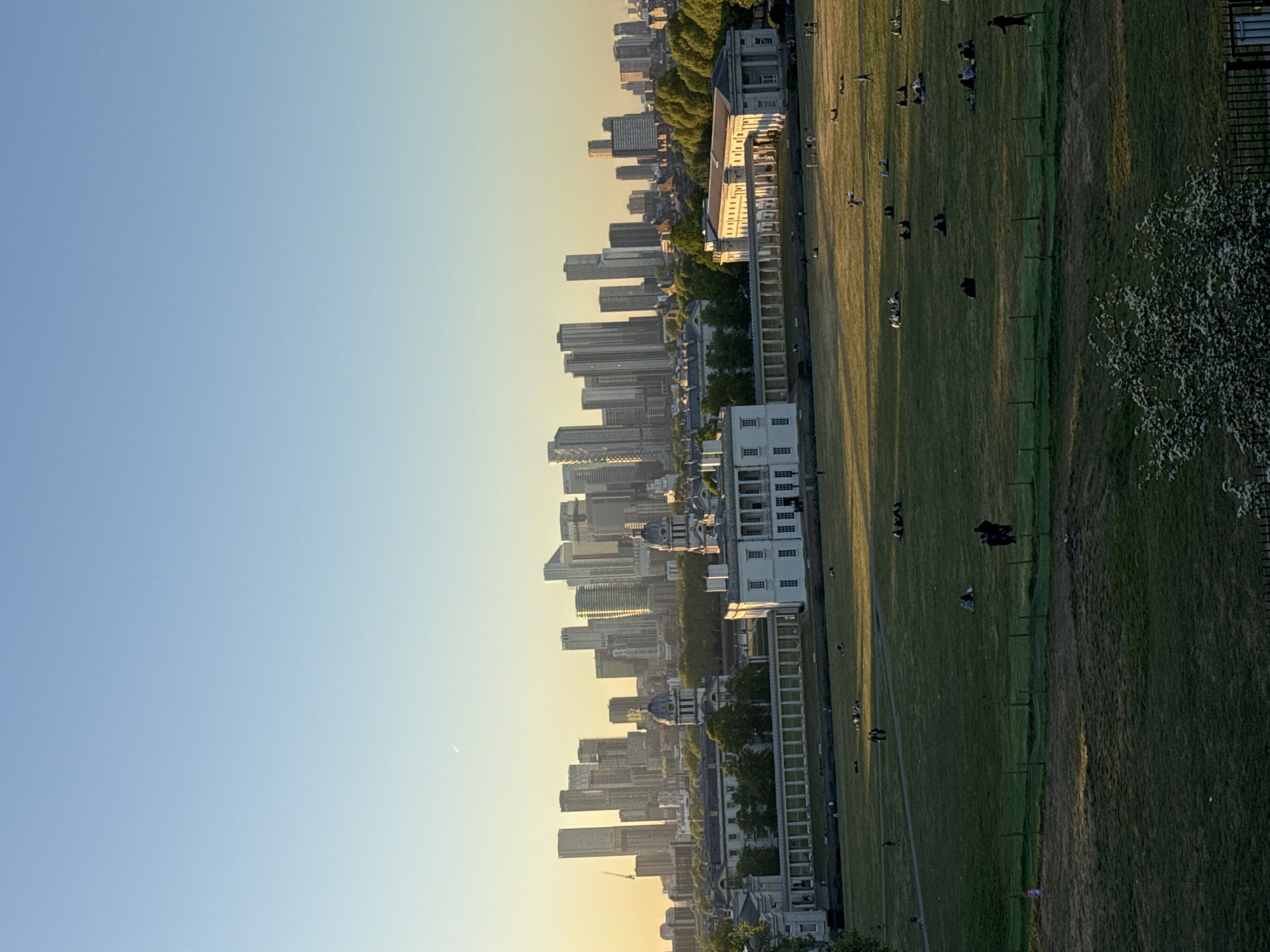 The image, when correctly oriented, presents a panoramic view of an urban landscape from what appears to be an elevated park during the golden hour.
In the foreground and midground on the right, a vast expanse of green grass is visible, indicative of a large park. Numerous individuals are scattered across the lawn, engaged in various leisurely activities such as walking, standing, or sitting. Some faint pathways traverse the grassy area. A blooming white-blossomed tree or bush is visible on the far right.
On the left side of the frame, a distinct cityscape unfolds. Closest to the park are several impressive classical white buildings, possibly institutional or historic, characterized by multiple windows and stately architecture. Beyond these, a dense collection of modern high-rise buildings and skyscrapers dominates the skyline, showcasing a contemporary urban development. The warm, low-angle sunlight illuminates their facades, creating a glow.
The sky is clear and features a gradient of colors, transitioning from a light blue at the top to a soft yellowish hue near the horizon, typical of either sunrise or sunset. A faint, thin white line, possibly a distant airplane contrail, is discernible in the upper part of the sky.
The scene captures a tranquil moment where people enjoy the open green space within a bustling metropolitan environment. The overall impression is one of urban leisure and harmony between nature and city life. Based on the architectural styles and the layout, particularly the combination of a park, historic buildings, and modern high-rises, the location is strongly suggested to be London, United Kingdom, likely from Greenwich Park looking towards areas like Canary Wharf. There is no visible text in the image.