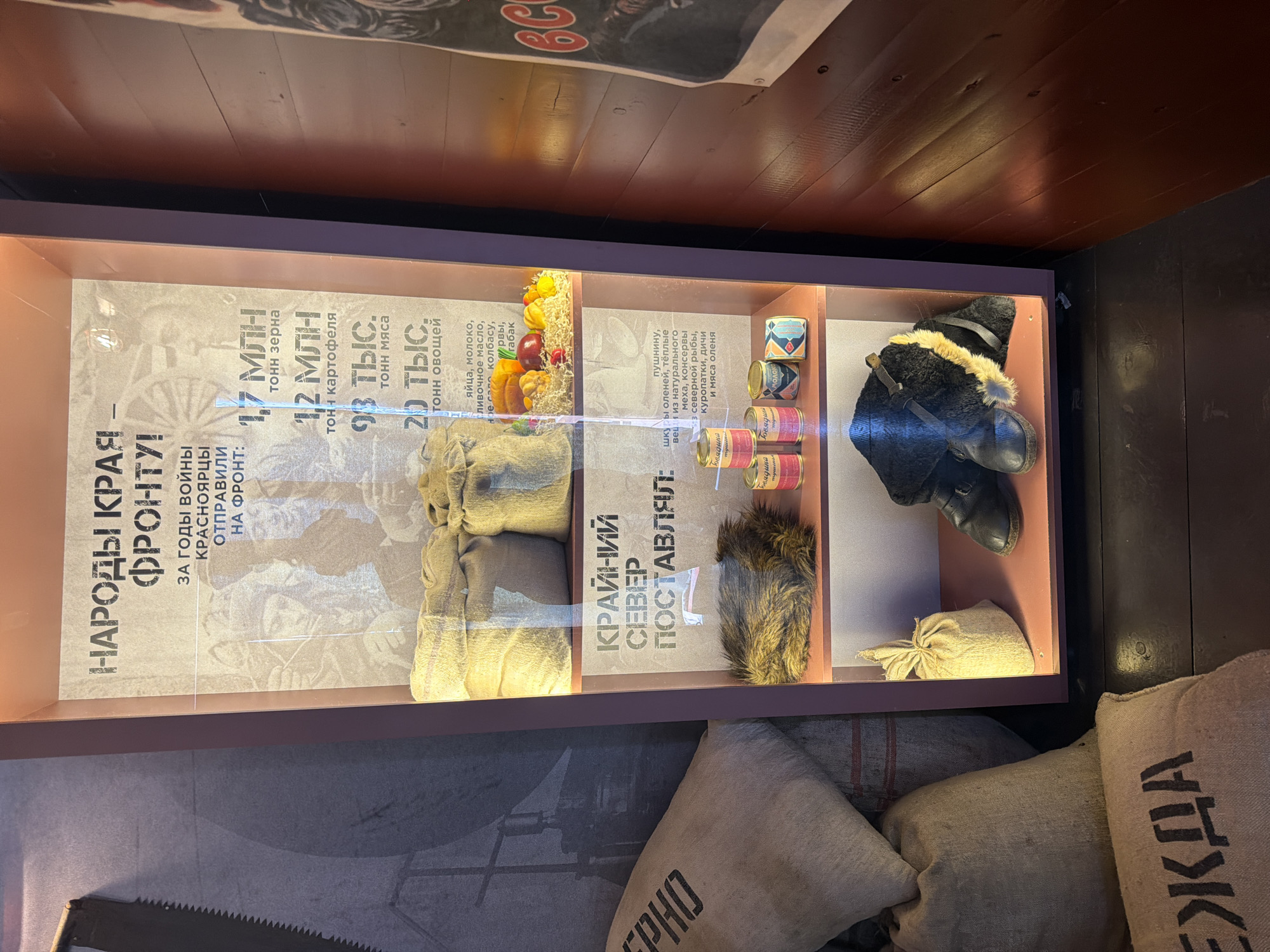 The image shows a display case with text in Russian, located in Krasnoyarsk, Russia. The text on the left panel translates to "The peoples of the region - to the front! During the war years, Krasnoyarsk sent to the front: 1.7 million tons of grain, 1.2 million tons of potatoes, 93,000 tons of meat, 20,000 tons of vegetables, eggs, milk, butter, sausage, tobacco, etc." The text on the right panel translates to "The Far North supplied: furs, reindeer hides, warm clothes made from natural fur, canned northern fish, partridge, game, and reindeer meat." In the display case are sacks, food items, canned goods, and a fur hat and boots. Below the display case are more sacks, one with Cyrillic text, possibly "Ezhda". The overall impression is of an exhibit depicting supplies and support for the war effort from the Krasnoyarsk region.