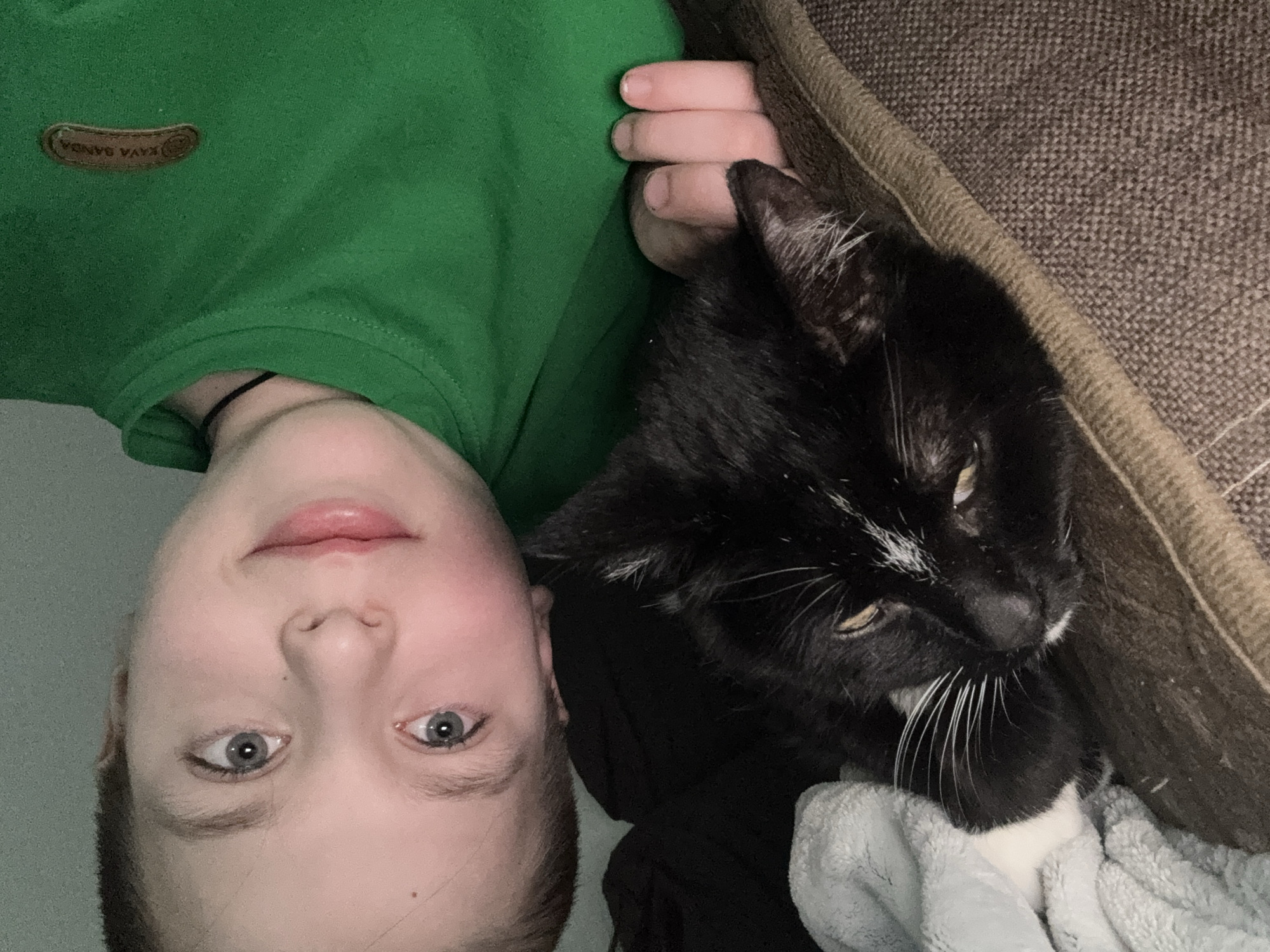 A person is lying down next to a black and white cat. The person has fair skin, blue eyes, and short blond hair. They are wearing a green shirt with a brown patch on the left shoulder. The cat is lying on a gray blanket next to a brown textured surface, possibly a couch or bed. The image is taken at an unusual angle, rotated approximately 180 degrees. The image does not contain enough information to determine a location in the city of Chaikovsky, Russia.