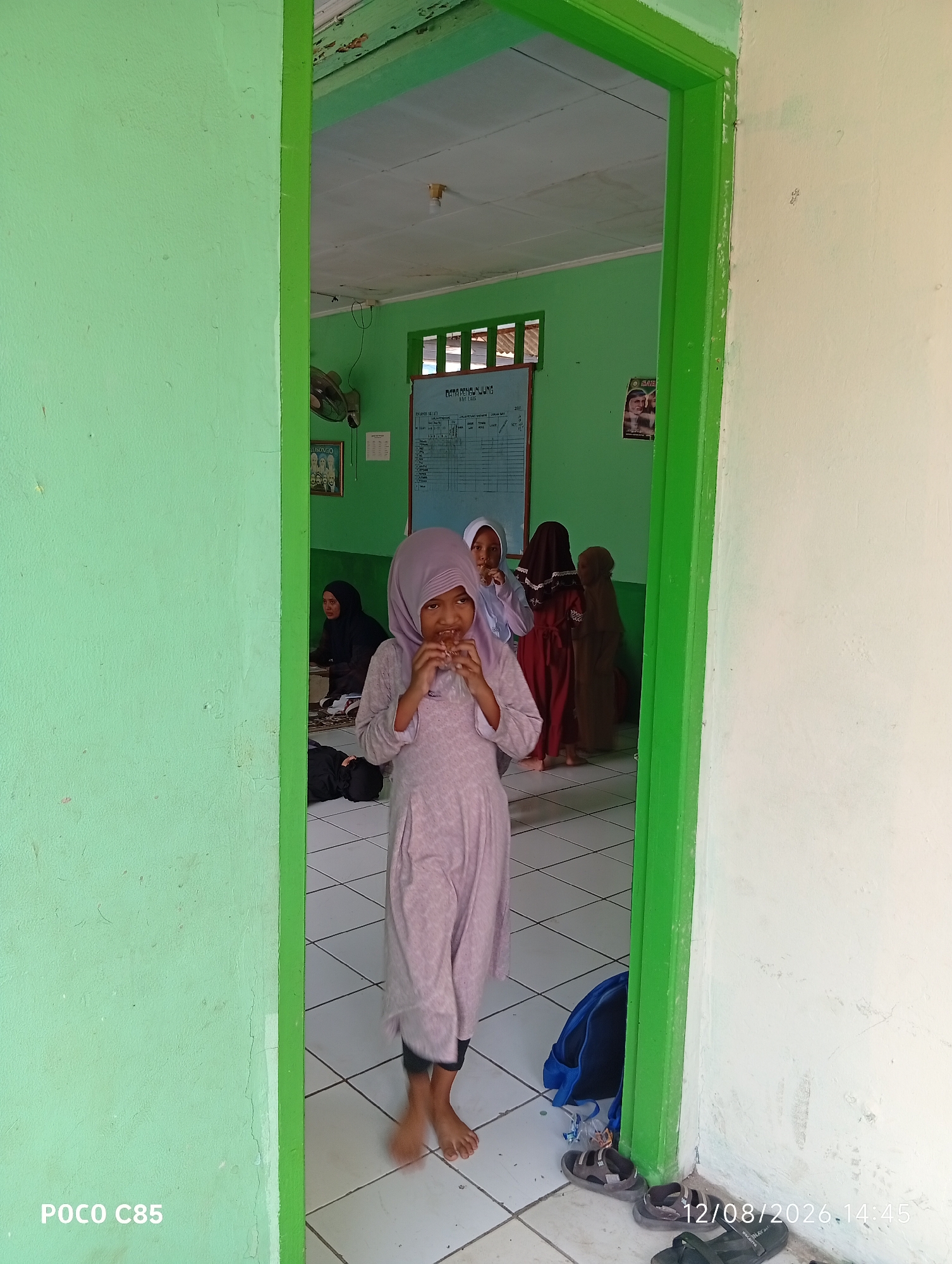 A young girl in a light purple hijab and matching long-sleeved dress stands in a doorway, holding something to her mouth. She has fair skin and dark hair visible beneath her hijab. Behind her, in a room with green walls and a tiled floor, several other children and adults are present. One woman in a black hijab and dress sits on the floor. Another child wears a dark brown hijab and dress, and a third wears a maroon dress and black hijab. A bulletin board with text and a framed picture are on the far wall. The doorway itself is painted bright green. The floor in the room is tiled in a diamond pattern. A blue backpack and several pairs of sandals are visible on the floor near the doorway. The timestamp "12/08/2026.14:45" is visible in the lower right corner. The phone model "POCO C85" is visible in the lower left corner. The image was taken in South Tangerang, Indonesia.
