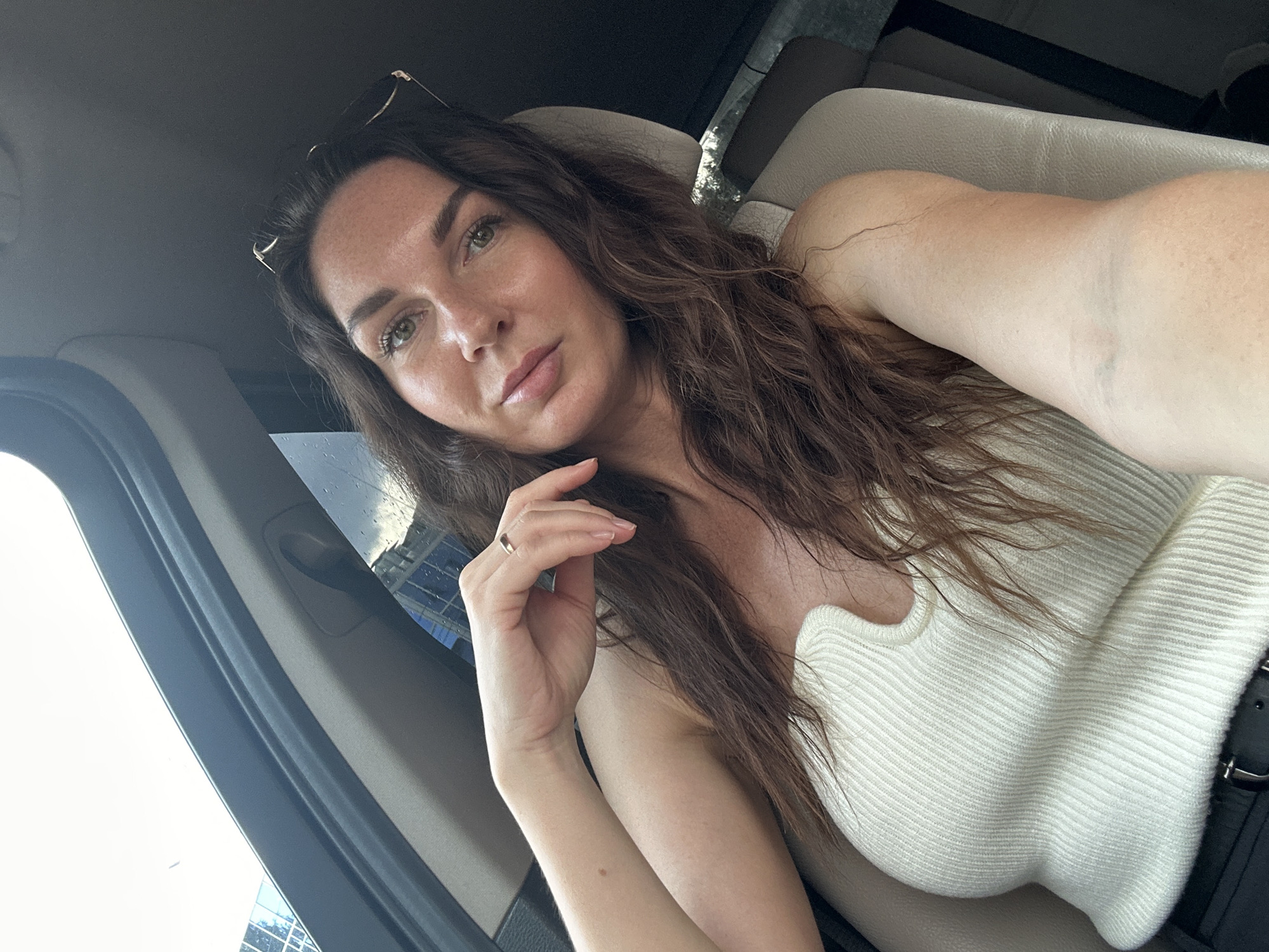 A selfie taken from the front passenger seat of a car shows a woman with long, wavy brown hair and green eyes. She is wearing a white ribbed tank top and a ring on her right hand. Sunglasses are perched on top of her head. The interior of the car, including the beige leather seat and gray trim, is visible. Outside the car windows, a modern building with blue glass and a cloudy sky can be seen, suggesting an urban setting. Based on the car's model and the surrounding architecture, this image was likely taken in the city of Chaikovsky, Russia.