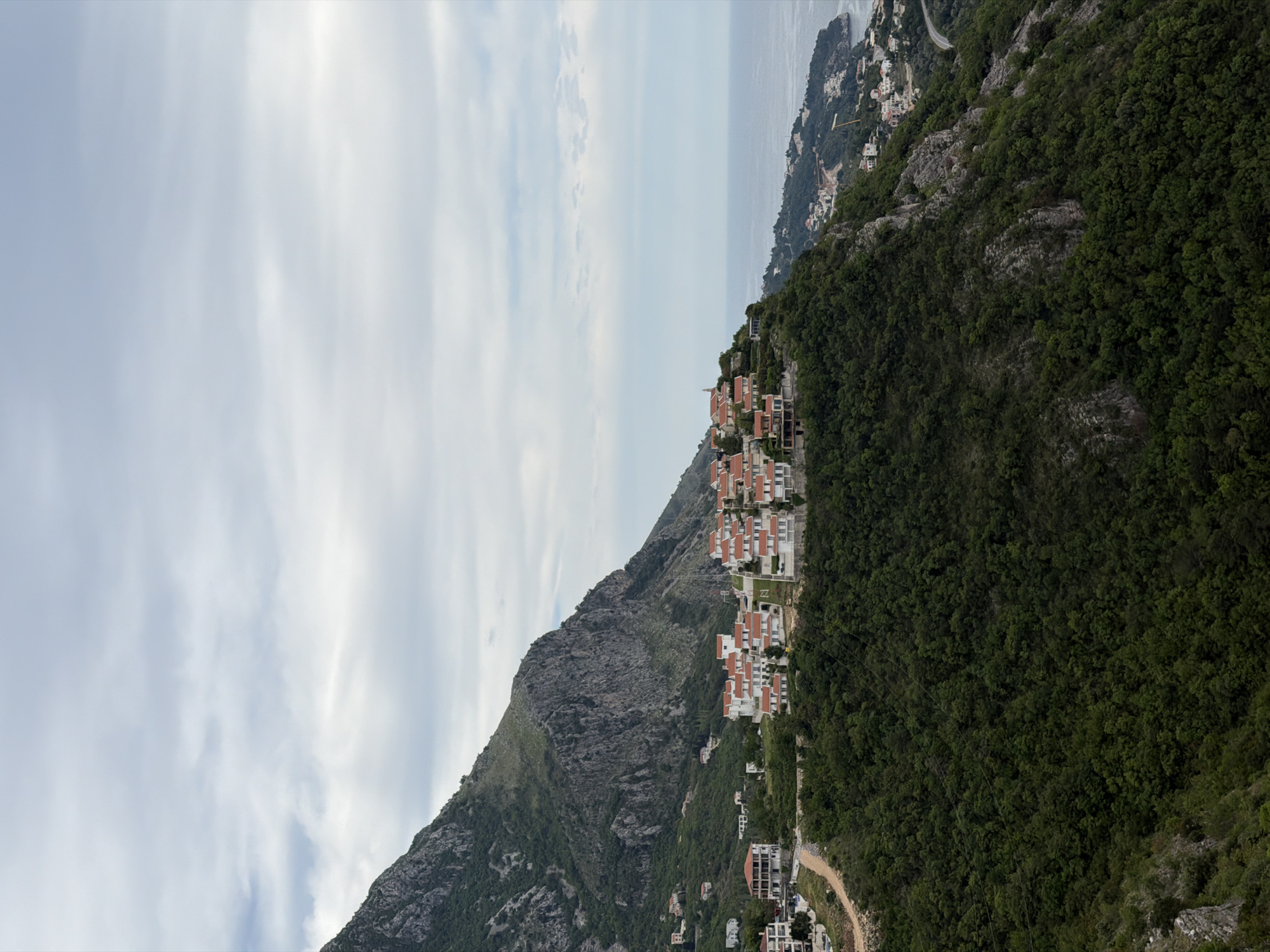 The image shows a hillside in Sveti Stefan, Montenegro, with buildings with orange tile roofs built along its side. The terrain varies from rocky to covered with dense green vegetation. A dirt road winds along the base of the hill. In the background, the Adriatic Sea is visible under a cloudy sky.