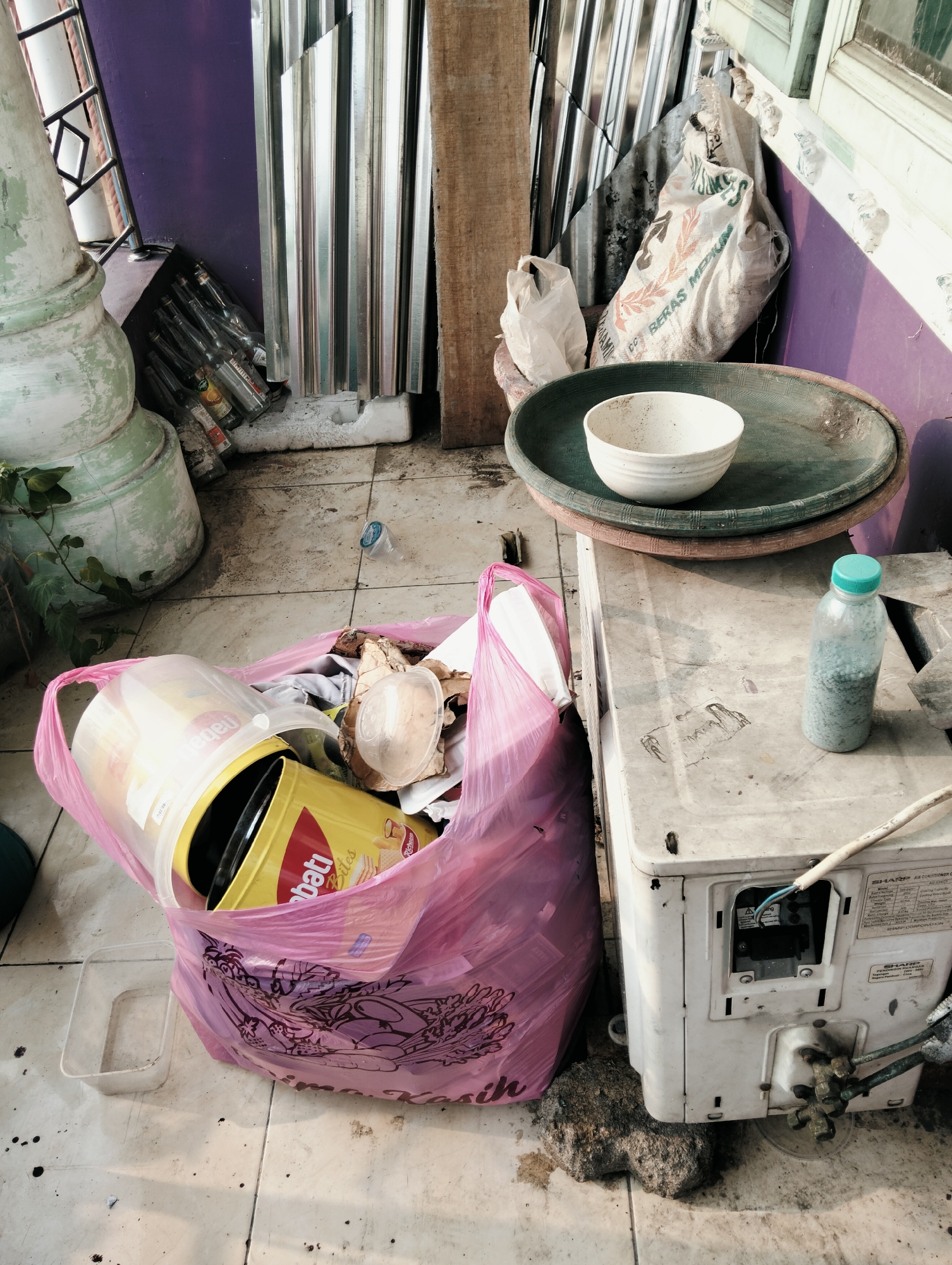 The image depicts a cluttered outdoor storage area, likely a balcony or alleyway, in the City of Medan, Indonesia. A large pink plastic bag overflowing with various items, including a yellow tin can with a red label, is prominently featured in the foreground. To the right, a white air conditioning unit sits on the tiled floor, supported by a rough rock. A plastic bottle filled with small blue granules and a green cap stands on top of the AC unit. In the background, stacks of corrugated metal sheets lean against a purple wall, alongside a large sack labeled "BERAS MEDIUM." Various glass bottles are visible near a large, pale green pillar on the left. The overall scene suggests disarray and neglect.