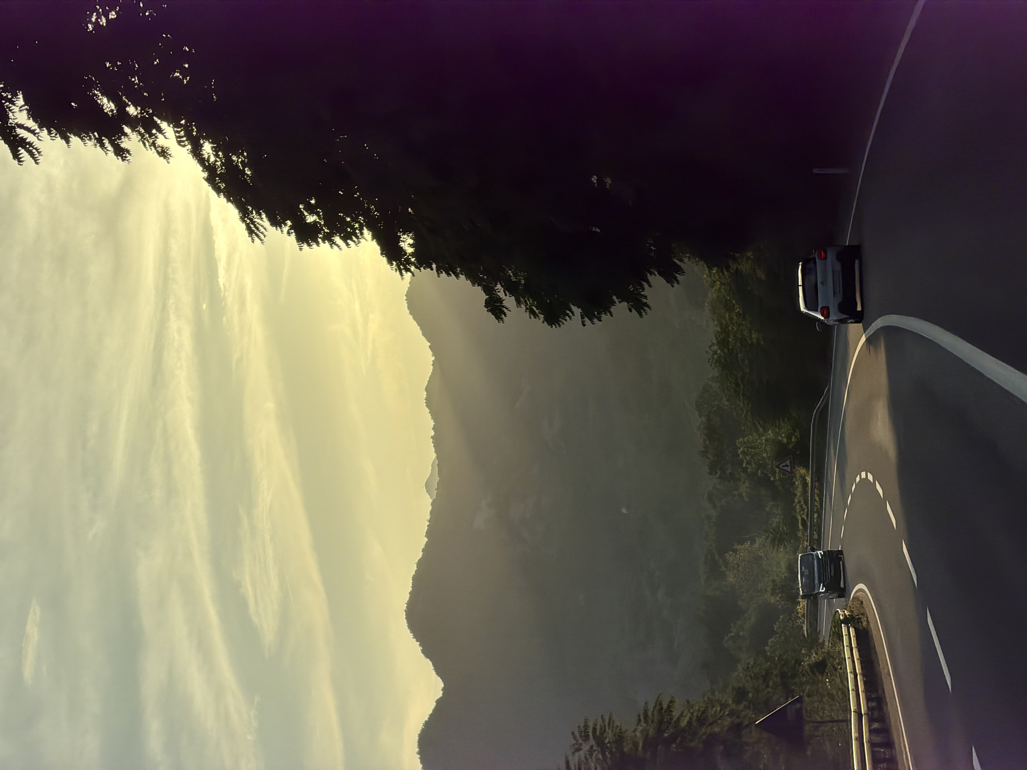The image shows a winding road in Gospoština, Montenegro, bordered by trees and a mountain range in the background. Two cars are visible on the road. The sky is light with scattered clouds.