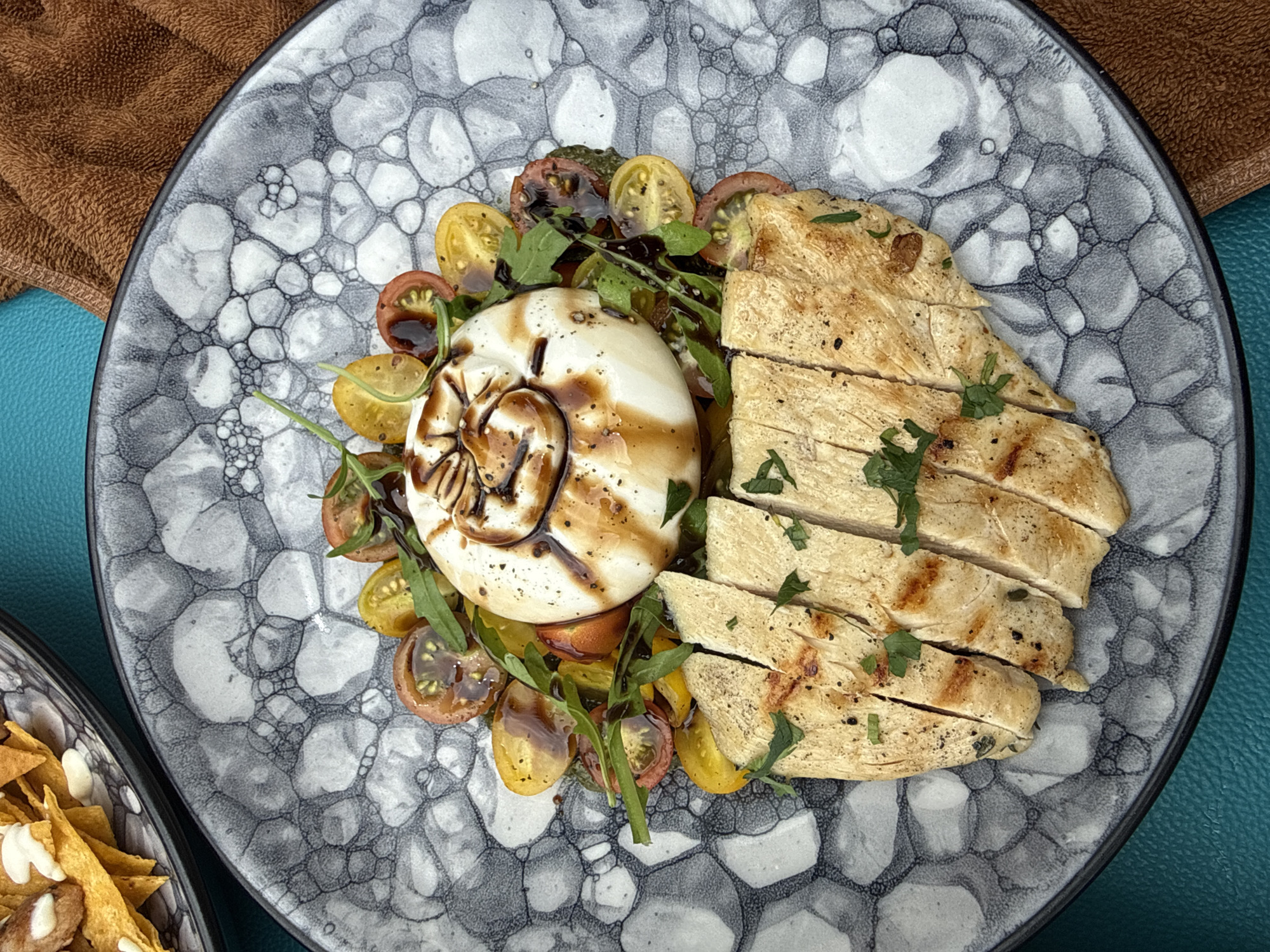 This overhead shot captures a vibrant and appealing meal served on a distinctive plate, likely enjoyed in Tegallalang, Indonesia. The main elements of the dish are sliced grilled chicken breast, a generous portion of cheese, and a fresh salad.
The grilled chicken breast is cut into several clean slices, exhibiting light grill marks and a succulent appearance. It is lightly seasoned and garnished with chopped green herbs, possibly parsley or cilantro, enhancing its freshness. Adjacent to the chicken is a large, round, white portion of cheese, likely burrata or mozzarella, distinguished by an artistic spiral drizzle of dark balsamic glaze or a similar reduction on its top surface.
Nestled beneath and around the chicken and cheese is a colorful salad. It features an array of halved cherry tomatoes in both rich red and bright yellow varieties, mixed with fresh green leafy vegetables, possibly arugula. The dark glaze from the cheese also lightly coats some of the salad components, tying the elements together visually.
The meal is presented on a striking grey ceramic plate that boasts a unique, organic, bubble-like, or cellular pattern in various shades of grey and white. This artistic plate adds a contemporary flair to the food presentation.
The background offers glimpses of the dining setting. A textured, warm brown fabric, possibly a napkin or part of a table covering, is visible in the upper left corner. A smooth, vibrant teal or blue-green surface, likely a tabletop or placemat, appears under the plate and to its right. Additionally, part of another dish containing golden, triangular chips topped with white dollops (suggesting nachos with sour cream or cheese) is partially visible in the bottom left, hinting at a wider meal spread.
The bright, even lighting suggests the photo was taken during the day. The overall impression is one of a thoughtfully prepared, fresh, and visually attractive meal. There is no visible text in the image.