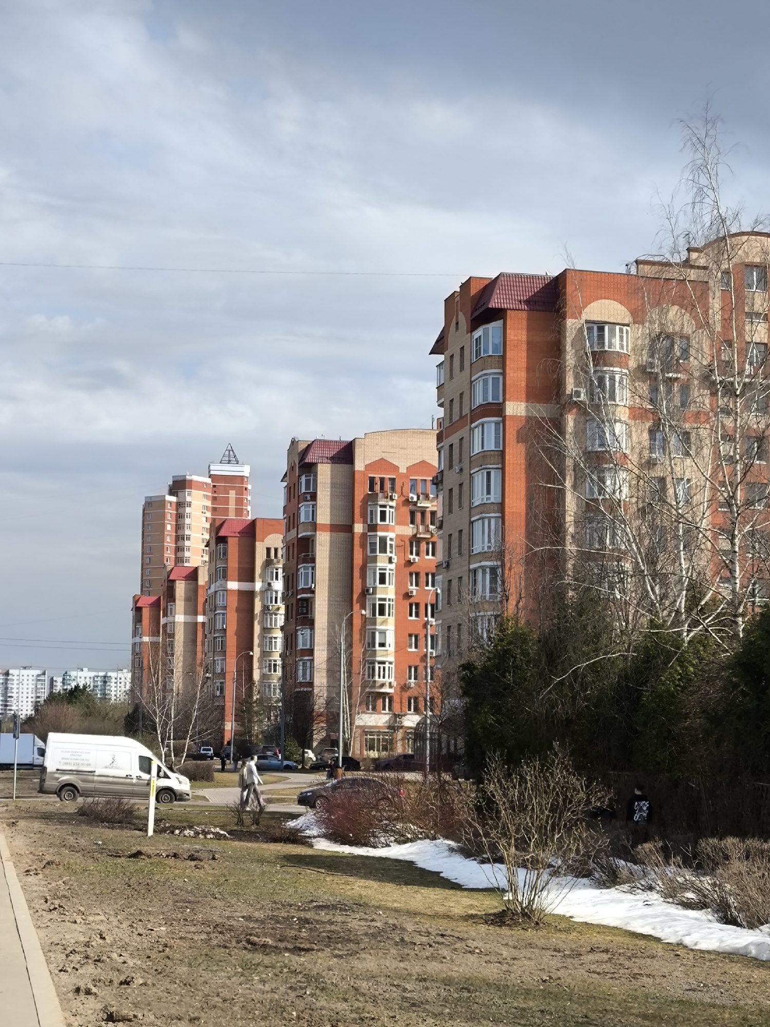 The image depicts a street in what is likely a residential area in Moscow, Russia. Several tall, red-brick apartment buildings with red roofs line the street. The buildings are of modern design, with many windows and some architectural embellishments.
In the foreground, there is a grassy area with patches of melting snow, suggesting it is early spring. Some bare trees and bushes are present. A white van is parked on the side of the road.
A person wearing light-colored clothing is walking along the street. Several parked cars are also visible. In the background, other buildings can be seen in the distance. The sky is overcast.