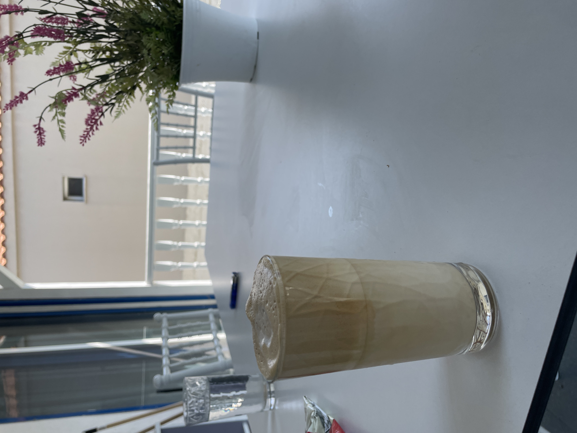 A tall glass of iced coffee sits on a white table outside a building in Skala Avdiron, Greece. The coffee is light brown and topped with a frothy layer. To the left of the coffee is a glass of water and a packet of something. Behind the table, there is a white railing and a building with a red-tiled roof. A potted plant with purple flowers sits to the right of the coffee.