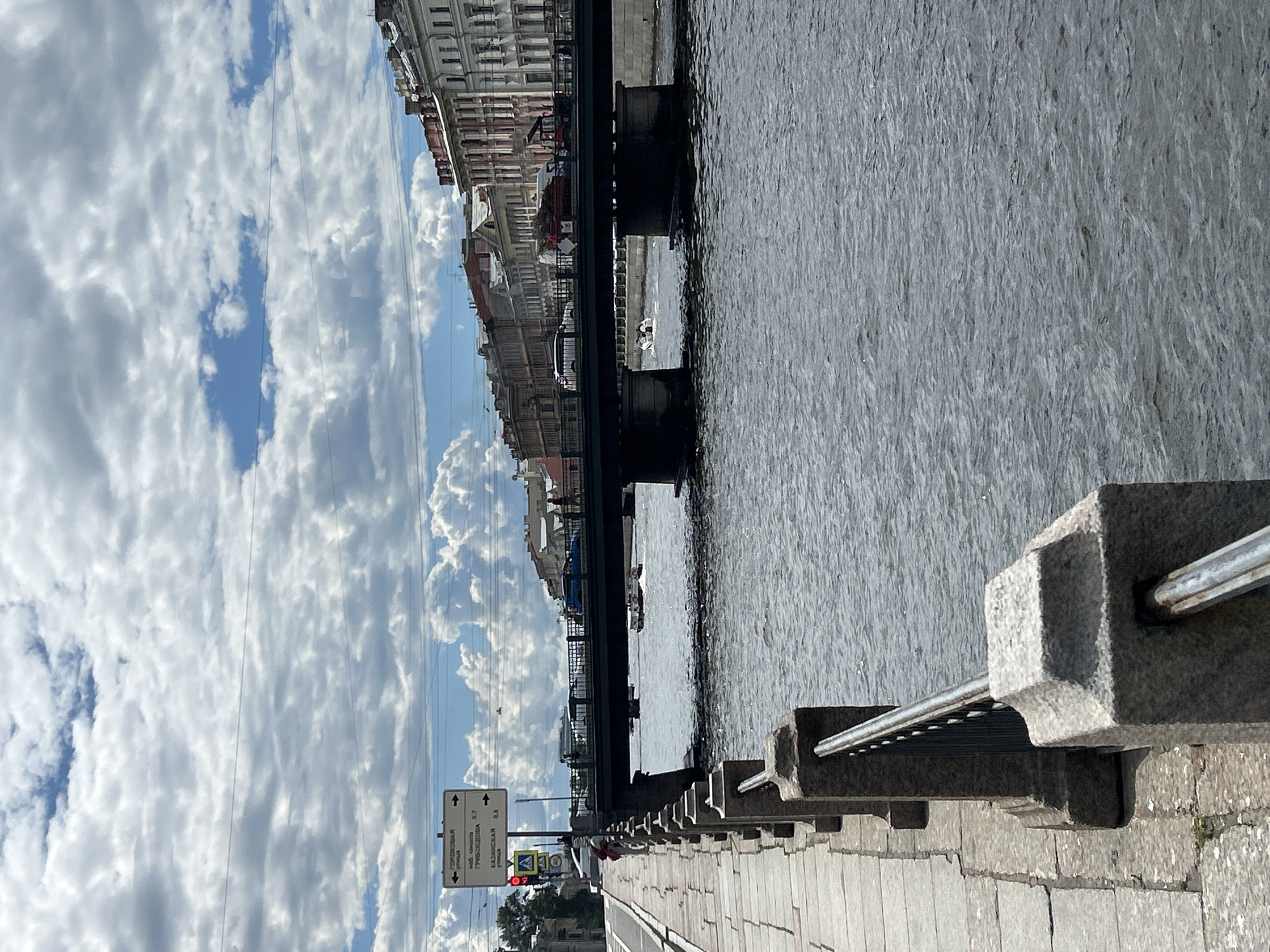 The image depicts a waterside scene with a bridge spanning across a river. On the left side of the image, a line of buildings with classical architecture lines the embankment. A pedestrian walkway with a stone railing and metal handrails is visible in the foreground, running parallel to the river. The water is choppy, suggesting a moderate breeze.
In the distance, under the bridge, a small white boat with two people is visible. The sky is filled with dramatic, puffy white clouds against a bright blue background, indicating a sunny day with variable cloud cover. A street sign with arrows and Russian text is positioned on the walkway, pointing towards "ТОРЖКОВСКАЯ улица" (Torzhkovskaya Street) and "ГРЕСНЕВИДНАЯ улица" (Gresnevidnaya Street), and "КАДАНДИНСКАЯ улица" (Kadandinskaya Street). A traffic light displaying a red signal is also visible near the signpost. Power lines traverse the sky. The overall impression is that of a pleasant day in an urban setting, likely a city with canals or rivers lined with historic buildings. The time of day appears to be daytime, likely midday given the bright sunlight and clear sky interspersed with clouds.