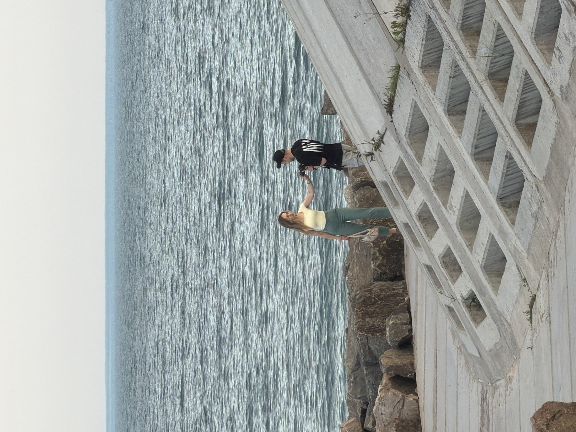 The image shows two people on a concrete embankment next to the sea. A woman with blonde hair, wearing a yellow top and green pants, stands on the left side of the frame, holding a light-colored purse. She is reaching out her left hand towards a man on her right. The man, wearing a black t-shirt and a black baseball cap, is holding a camera and appears to be taking a photo of the woman. The sea is visible in the background, with a calm surface and a faint horizon. The embankment is made of large, rough stones and a stepped concrete structure with square openings. The overall scene appears to be outdoors, likely in a coastal area. The location is identified as Fontanka, Ukraine.