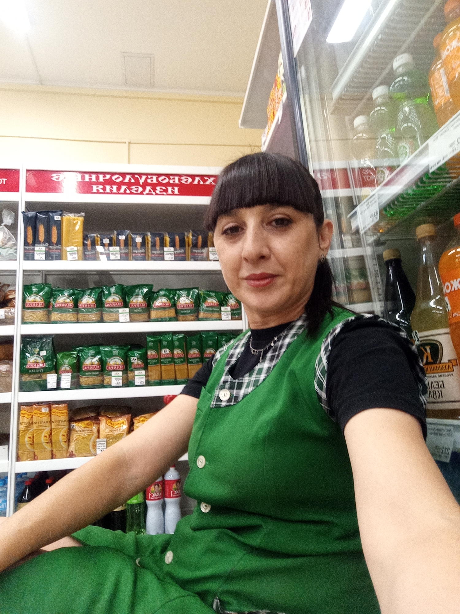 In a brightly lit retail environment, a woman with dark, bobbed hair and bangs, wearing a black shirt under a green plaid vest, poses for a selfie. She is positioned in front of shelves stocked with various food products, including pasta and bottled beverages. To her right, a refrigerated display case contains more drinks. The setting appears to be a grocery store or small market, likely in the city of Belogorsk, Russia, given the Cyrillic text visible on signage and product packaging. The overall impression is a candid moment captured by an employee during her workday.