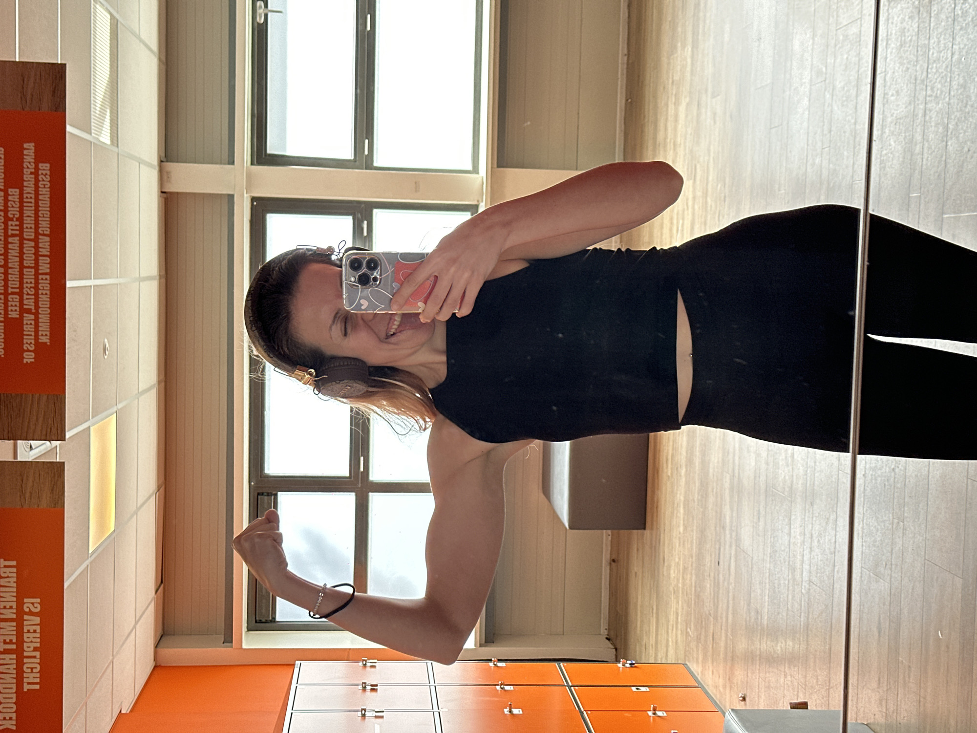 A woman is taking a mirror selfie in what appears to be a locker room. She is wearing black athletic wear and headphones. She is flexing her bicep. The room has lockers, mirrors, and a wooden floor. There is text on the wall that appears to be in Dutch.