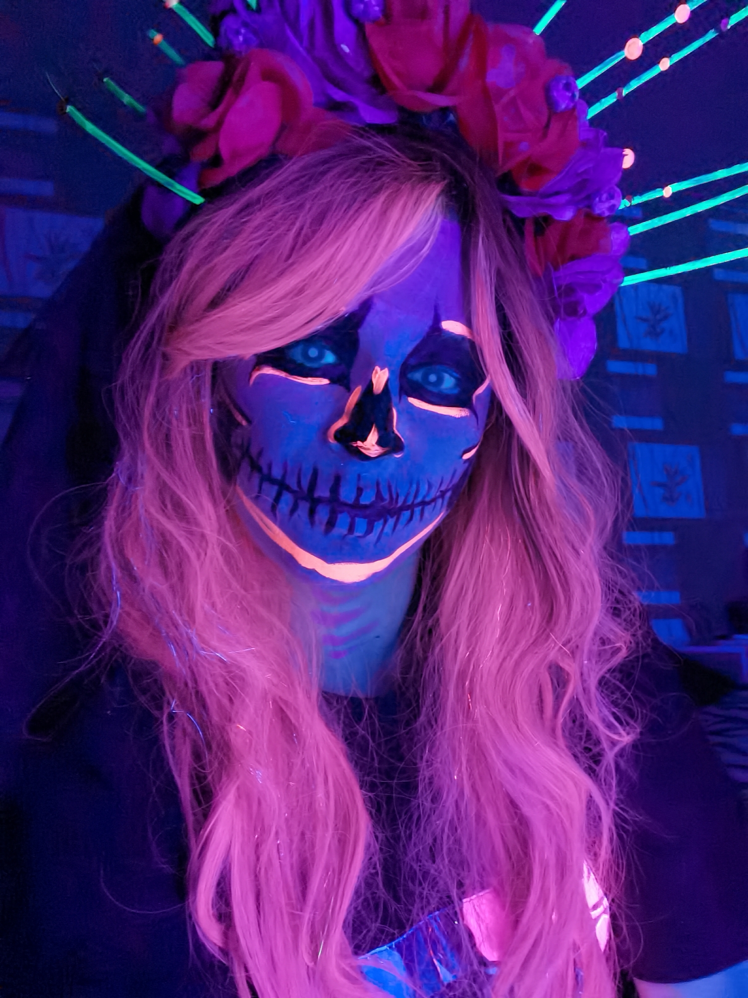 An indoor portrait taken in Chaikovsky, Russia, features a person with long, wavy, light pink hair, brightly illuminated by ultraviolet (UV) light. The subject's face is meticulously painted with UV-reactive makeup, forming a glowing sugar skull design. The base of the face paint is a deep purple-blue, while the skull's features—such as the prominent eye sockets, a stylized nose, and stitches across the mouth—are rendered in vivid, neon orange. The person's natural blue eyes are striking against the painted face.
She wears a decorative floral crown or headband, adorned with purple and red flowers. Extending from the crown are several thin, glowing neon-green rods that radiate upwards, contributing to a distinctive halo effect. The individual appears to be dressed in a dark, possibly black, top.
The background is dark, characteristic of a UV-lit environment. It features several glowing neon-green horizontal lines, some punctuated with small, glowing orange dots, creating a decorative light installation. Below these, square patterns, likely decorative panels or framed art, are faintly visible with a blue luminescence on the wall. The scene evokes a vibrant and artistic atmosphere, suggesting a themed event, party, or celebration, such as Halloween or Día de los Muertos, where special effects makeup and blacklight are key elements. The person maintains a neutral expression, allowing the intricate and glowing makeup to dominate the visual narrative. No visible text is present in the image.