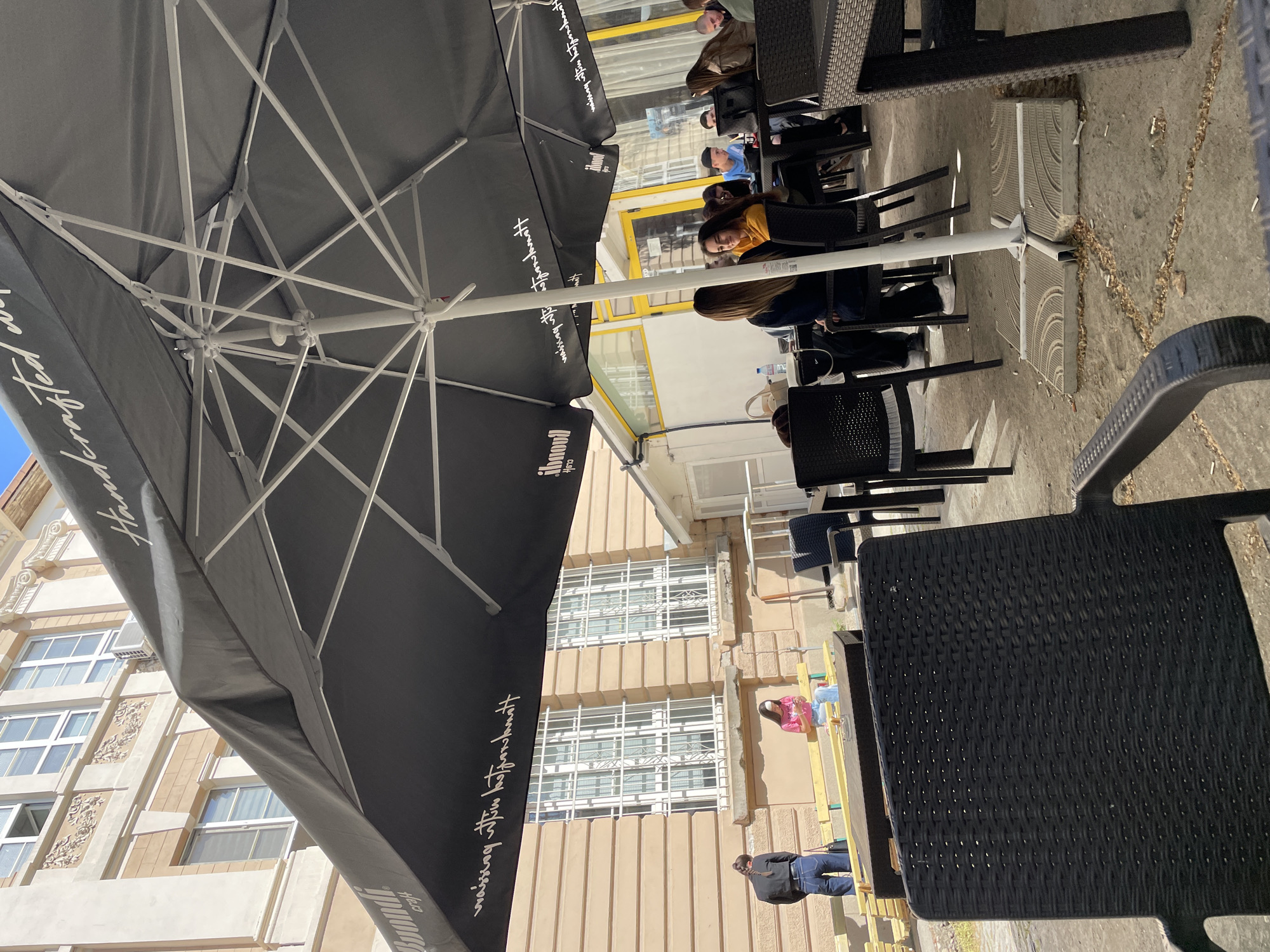 The image captures an outdoor cafe scene in Plovdiv, Bulgaria, on a bright, sunny day. The perspective is taken from under a large, dark-colored market umbrella, which dominates the left foreground and features a white metal frame. The umbrella has white text printed on its fabric, including "Handcrafted" and what appears to be stylized Asian script and the word "Havana" and "il mondo" or similar branding.
Several dark-colored, wicker-style tables and chairs are arranged on a concrete or paved patio area. Numerous patrons are seated at these tables. Notable individuals include a person with long brown hair wearing a yellow top, a person in a blue top with a baseball cap, and another person in a brown jacket, all appearing to be relaxing or socializing. On one table, a water bottle is visible.
In the background, two buildings line what appears to be a street or alleyway. The main building features a light beige or cream-colored facade with decorative architectural elements and multiple windows, some fitted with white security grates. An air conditioning unit is visible on this building. Another section of a building has large, yellow-framed windows.
Near the buildings, a person in a dark jacket and jeans stands, and another person in a pink top is seated on what looks like a temporary wooden structure or low platform. The ground shows some scattered debris, including leaves and what appear to be cigarette butts, especially around the base of the umbrella, which is secured by heavy concrete blocks with a patterned surface. The overall atmosphere suggests a casual outdoor dining or social setting.