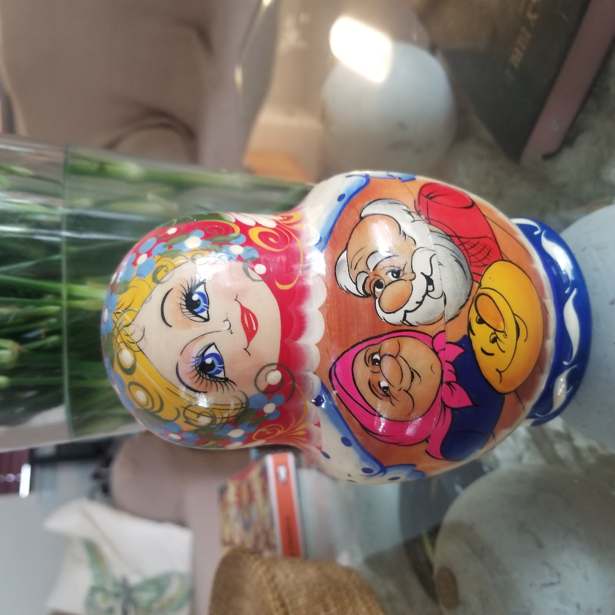 A matryoshka doll is displayed on a glass table, with a blurred background showing a couch and a vase of flowers. The doll is intricately painted with a woman's face on the top, featuring blue eyes, red lips, and blonde hair. Below her face, there are two smaller figures: a woman with a pink headscarf and a man with a white beard. A yellow oval shape with a smiley face is painted at the bottom of the doll. The doll has a blue and white patterned base. The scene is likely indoors, possibly in a residential setting in Virginia Beach, United States of America.