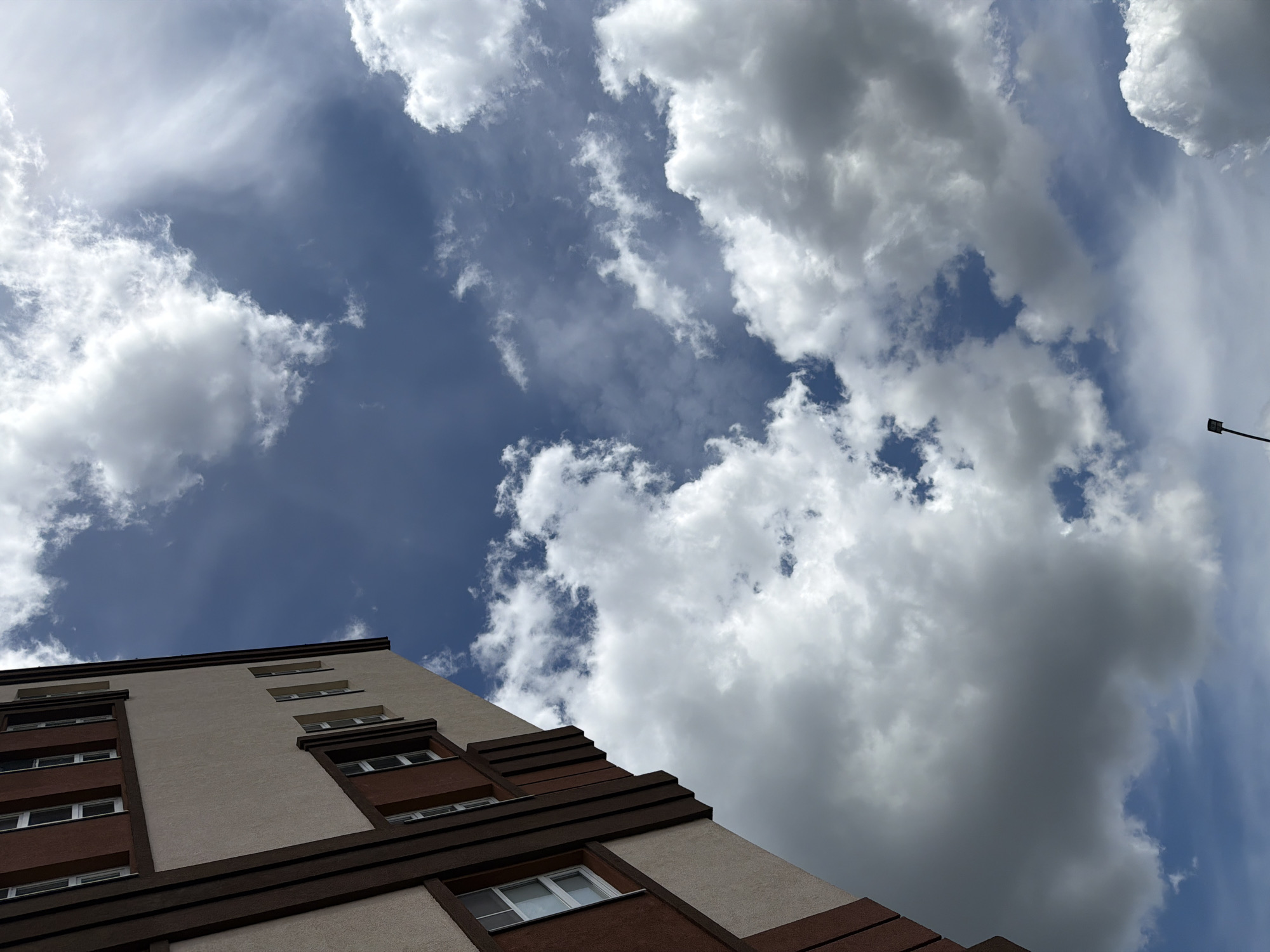 This is a photograph of a building exterior against a partly cloudy sky, taken from a low angle looking upwards. The building is modern, with light beige siding and dark brown trim framing rectangular windows. The windows are white and appear to be double-paned. The sky is a mix of blue and white clouds, with some areas of darker, heavier clouds and lighter, wispy ones. The photograph is taken in Lopatino, Russia, and the light suggests it is daytime. There are no people visible in the frame.