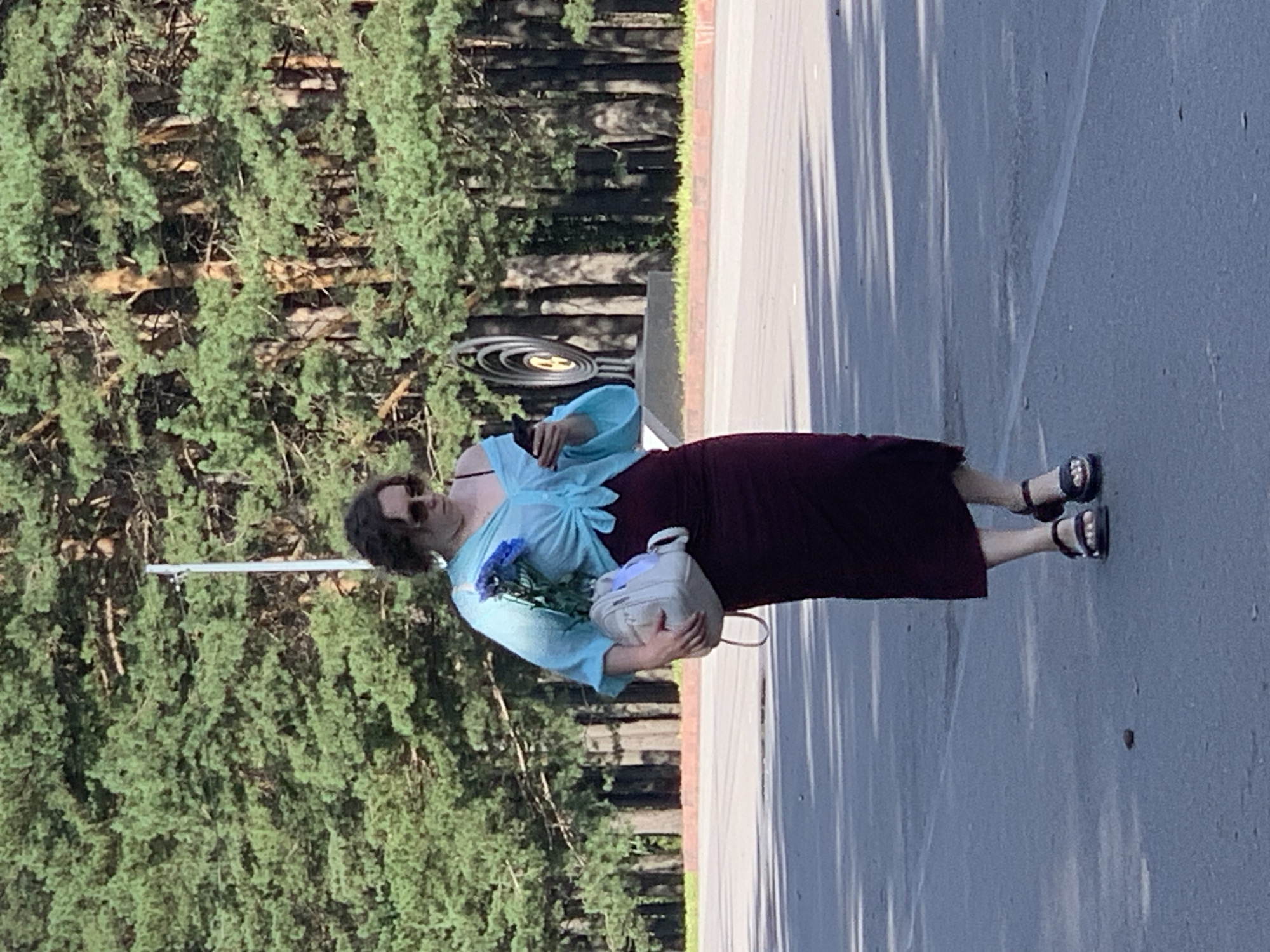 The image shows a woman walking outdoors on a paved surface, possibly a road or sidewalk. She is carrying a light-colored backpack in her left arm and a bunch of blue flowers in her right hand. She is wearing a light blue, off-the-shoulder top tied at the waist, a dark maroon skirt that reaches her ankles, and dark sandals. She is also wearing sunglasses and appears to be looking at her mobile phone, which she holds in her right hand. Behind her, there is a dense line of tall green trees, likely pines, forming a natural backdrop. In the middle ground, to the right of the woman, a distinctive metal sculpture with concentric circles is visible. The ground in the foreground is asphalt, with some scattered debris. The lighting suggests it is daytime, with sunlight filtering through the trees, creating dappled shadows on the ground. The image quality is moderate, with some slight blurriness. The specific location in the city of Prokopyevsk, Russia, is not identifiable from the visual elements in the image.