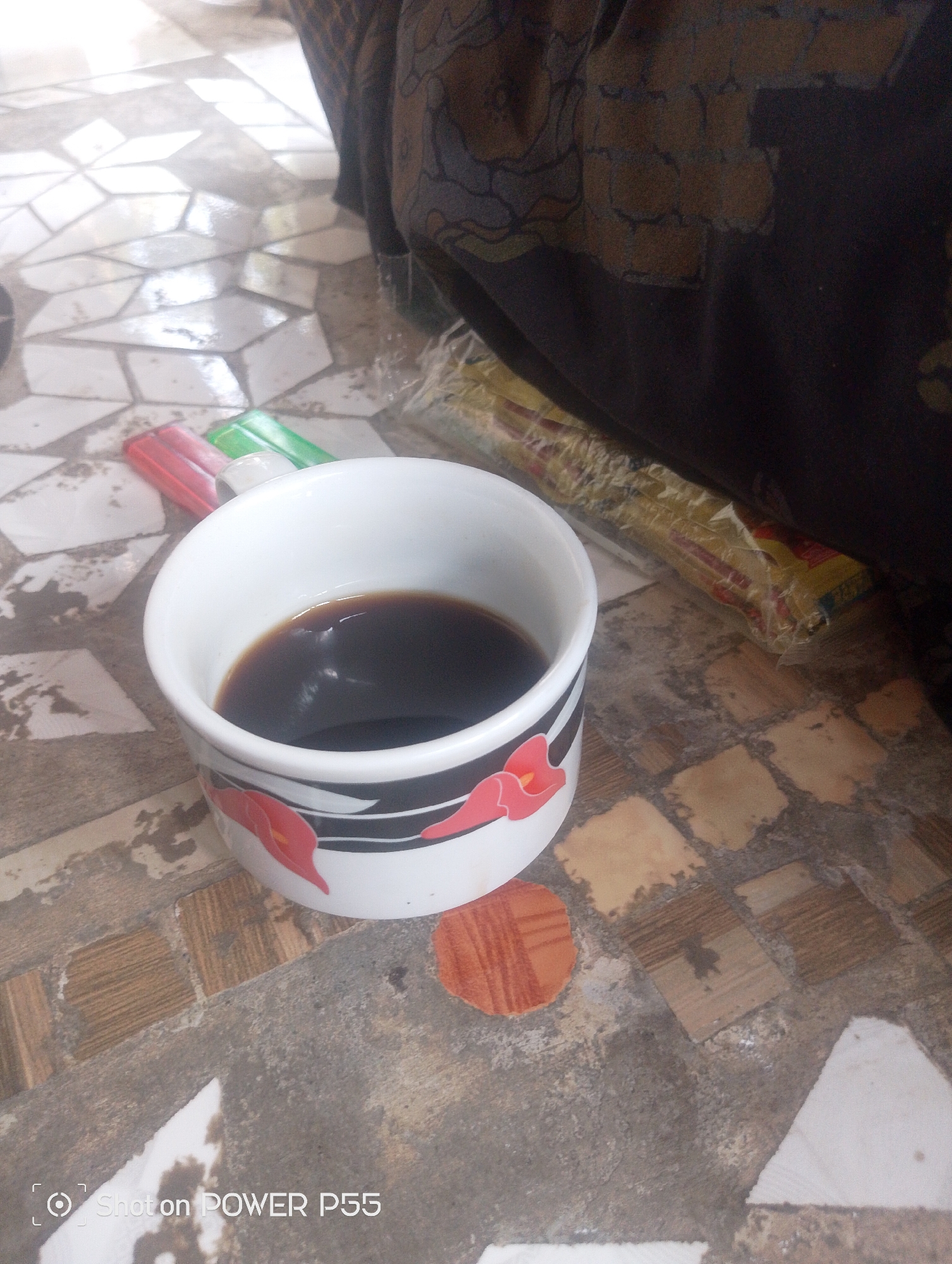 A white ceramic mug with a floral pattern containing a dark liquid, likely coffee, is placed on a floor composed of various irregularly shaped tiles. To the left of the mug are two red and green objects, possibly markers or pens. Behind the mug, a portion of a dark fabric with a patterned design is visible, and to the right of that, a long, rectangular package wrapped in clear plastic with yellow and red labeling. The floor tiles exhibit a mosaic-like appearance with variations in color and texture, including some lighter, reflective areas. The scene is lit, casting subtle shadows. The image includes a watermark at the bottom left corner reading "Shot on POWER P55". This scene is likely situated in an indoor setting in Lebaksiu, Indonesia.