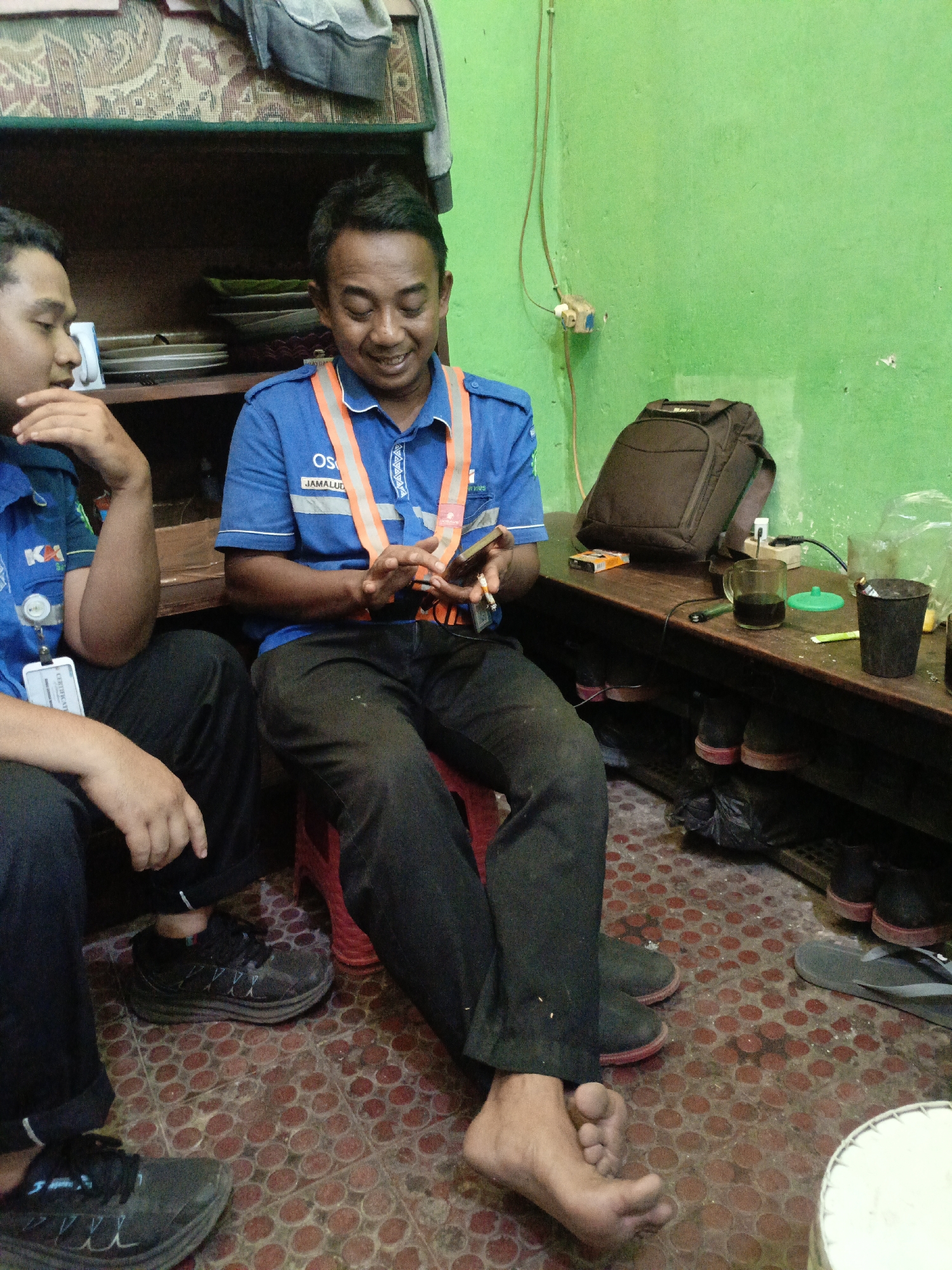 The image depicts two men in the city of Kidul Dalem, Indonesia, likely in a workshop or break room. The man on the right, wearing a blue uniform with an orange safety vest, is smiling and looking down at a mobile phone in his hands. He appears to be holding a cigarette. Next to him, a backpack rests on a wooden shelf that also holds a glass of dark liquid, a pack of cigarettes, and other miscellaneous items. The shelf is stocked with shoes underneath. The man on the left, also in a blue uniform, is partially visible and appears to be talking to the other man. The room has a green painted wall and a tiled floor with a red circular pattern. The overall environment suggests a casual and utilitarian setting.