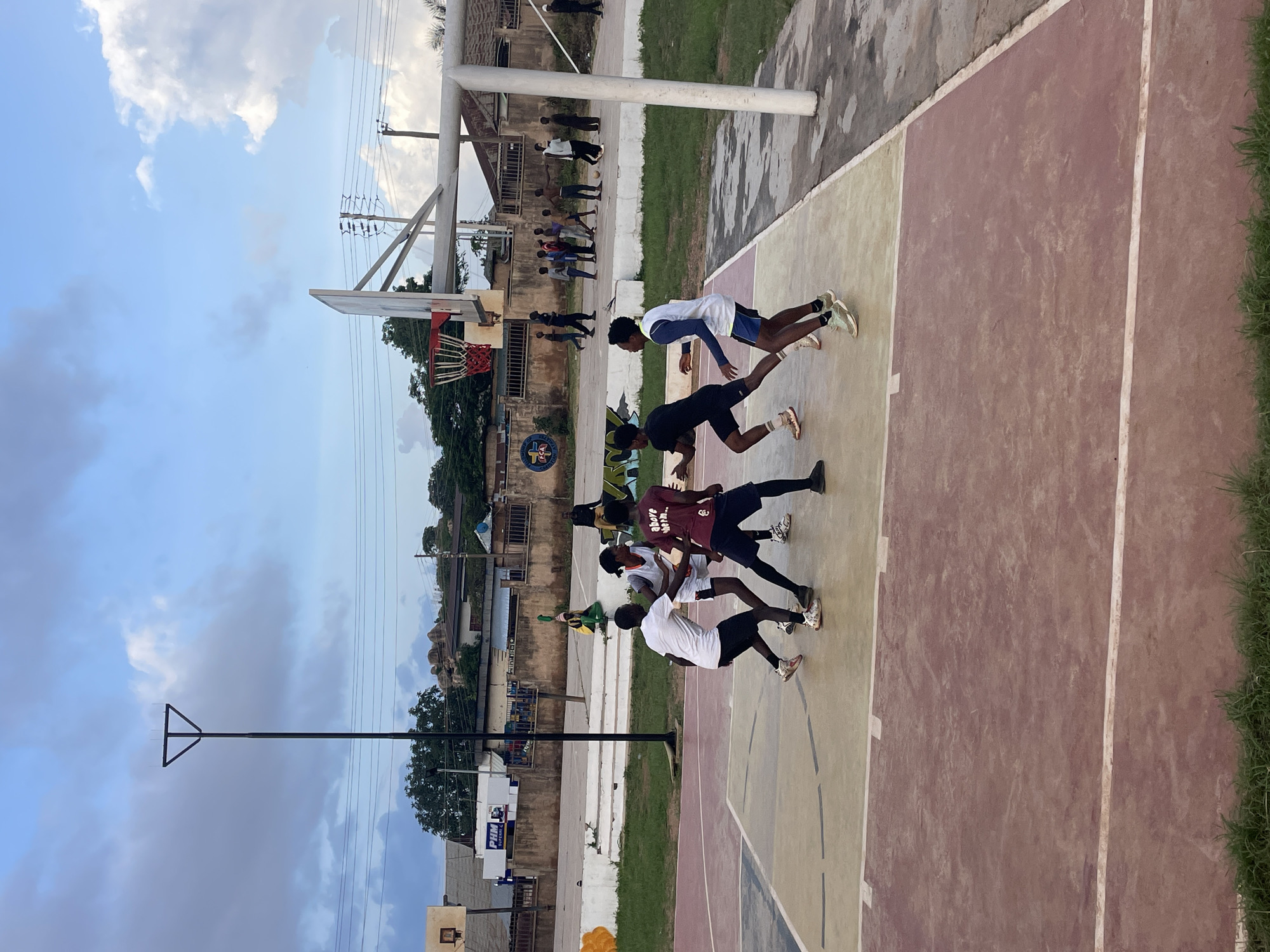 The image depicts a group of young men engaged in a basketball training session on an outdoor court. The court surface is a mix of reddish-brown and beige concrete, with white lines marking boundaries. A basketball hoop with a red net is visible. In the background, there are buildings, some appearing to be residential or shops, with corrugated metal roofs. Trees and vegetation surround the court. Overhead, a bright sky with scattered clouds suggests it is daytime, likely late afternoon or early evening given the soft lighting. The individuals are dressed in athletic wear, and their postures indicate they are actively participating in drills or practice. One person in the foreground is wearing a white t-shirt and black shorts, another in a maroon shirt and black shorts, and another in a black t-shirt and shorts. Several other figures are further back on the court. The overall mood appears to be energetic and focused. One of the buildings in the background has text that reads "PHM" on a sign. The location is Gura Top, Nigeria.