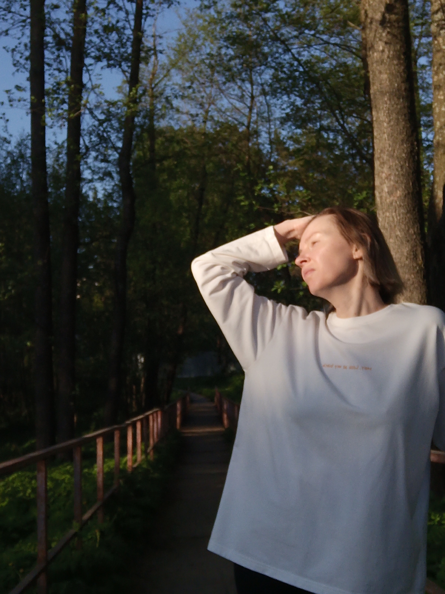 A woman stands on a path in a wooded area, likely in a park or forest in or near the city of Chaikovsky, Russia. She is wearing a white, long-sleeved shirt and dark pants. Her right hand is raised and resting on her forehead. The path is bordered by a low, rusted railing. Trees surround the path, creating a backdrop of dense foliage. The lighting suggests it is either early morning or late afternoon, with some areas illuminated by direct sunlight and others in shadow.