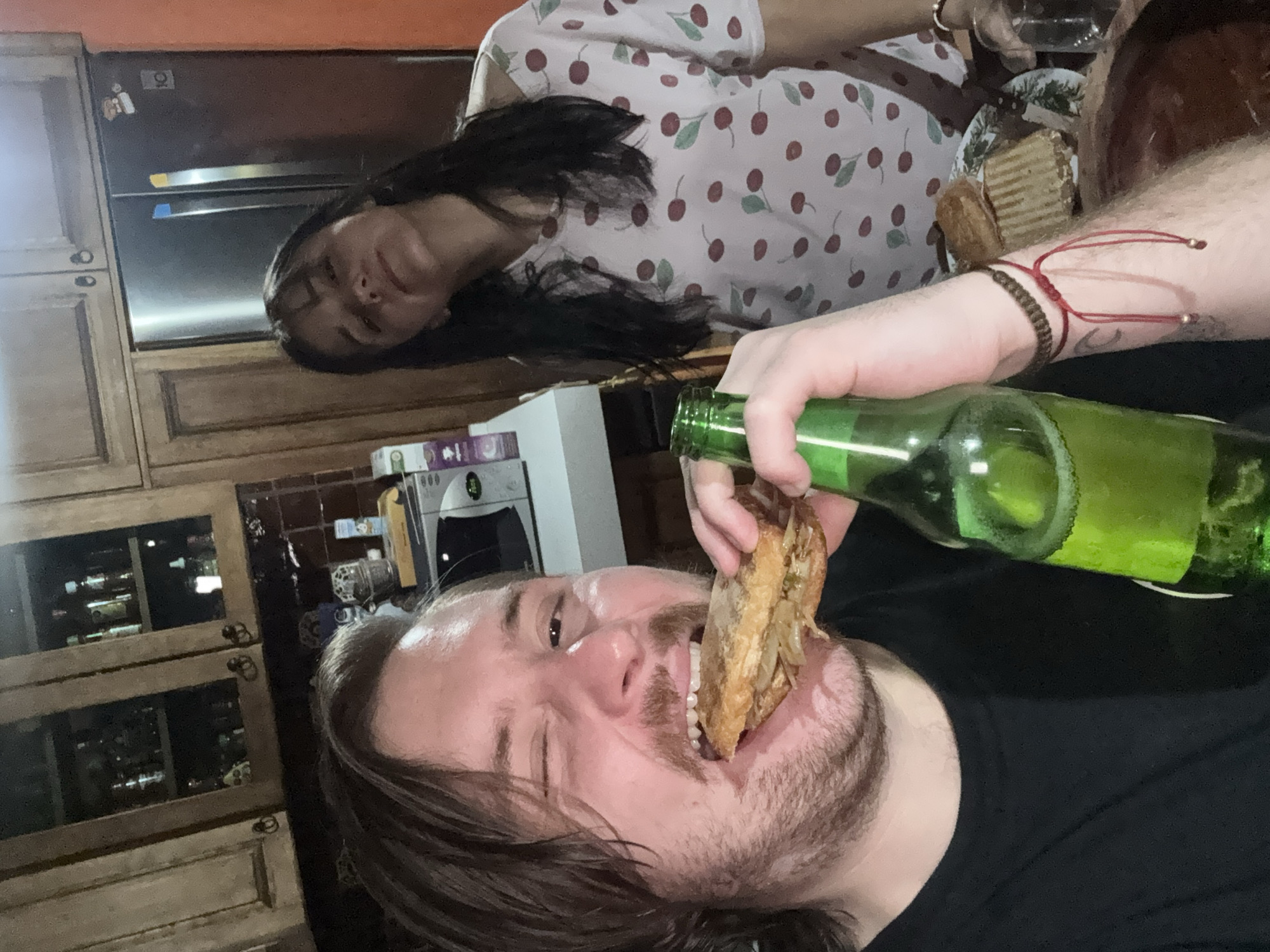 The image shows a man with brown hair and a beard, holding a bottle of green beer and taking a large bite of a sandwich. His arm has two bracelets. To the right, a woman with long dark hair is wearing a cherry-patterned shirt. In the background, there is a kitchen with wooden cabinets, a refrigerator, and a microwave. The image was taken indoors. There is no information to establish the location in the city of Cañada de Madero, Mexico.