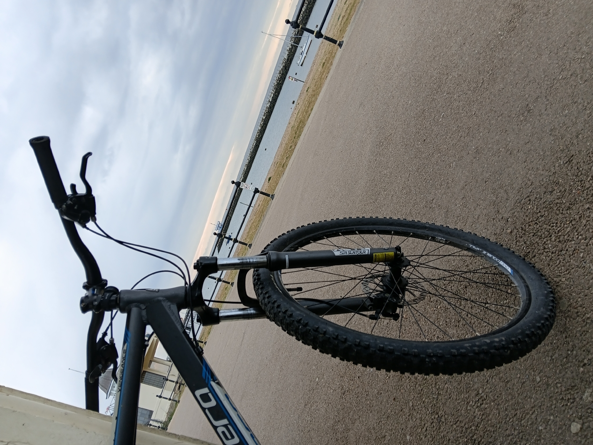The image depicts a bicycle on a paved surface, likely a path. The bicycle is a mountain bike, with a black frame and blue accents. The front wheel is prominently visible, with a knobby tire. The handlebars and front fork are also in view. In the background, there is a body of water with a breakwater or pier-like structure extending into it. There are posts along the edge of the water. Further in the distance, there are boats. The sky is overcast. Based on the visual information, a precise location in Canterbury cannot be accurately determined. The scene appears to be a coastal or waterfront area.