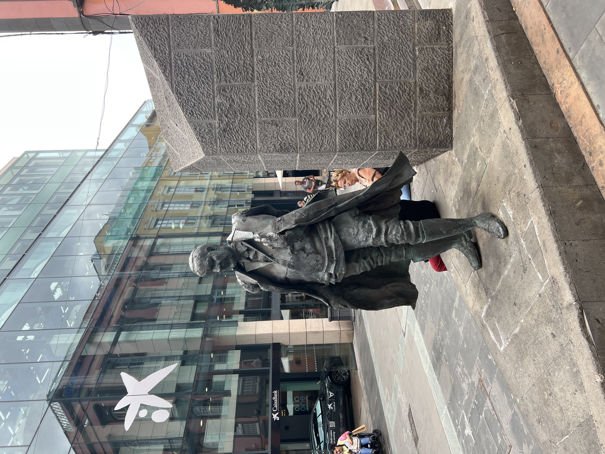 The image shows a bronze statue of a man standing on a stone base in front of a modern glass building. The building has a logo of CaixaBank on it, indicating it's a bank branch. The statue is in Las Palmas de Gran Canaria, Spain. People can be seen in the background, including a woman leaning on the stone structure behind the statue. A black car is parked outside the bank and a person in a wheelchair is also present.