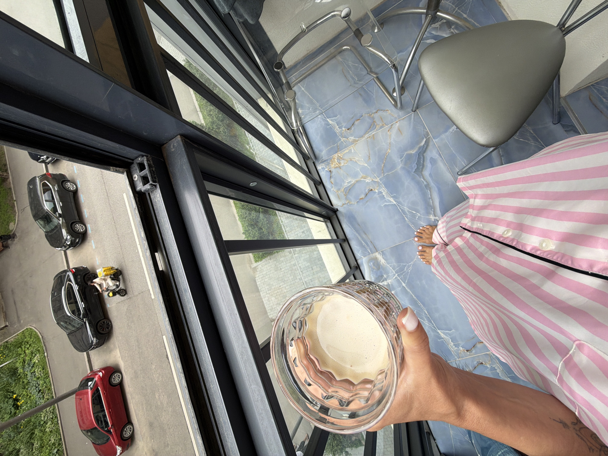 In a high-angle shot, a person wearing pink and white striped pajamas stands on a balcony with a blue marble tiled floor. Their feet, with white-painted toenails, are visible. The person is holding a clear glass filled with a light-colored beverage, possibly coffee or a smoothie. To the left of the frame, a large glass window looks out onto a street with several parked cars, including a red sedan and two black cars. A person is seated on a small motorized vehicle, possibly a scooter, between the two black cars. The scene appears to be set during the daytime, with natural light illuminating the area. The presence of cars and a building suggests an urban environment, consistent with the provided location context of Moscow, Russia. There are no visible text elements in the image.