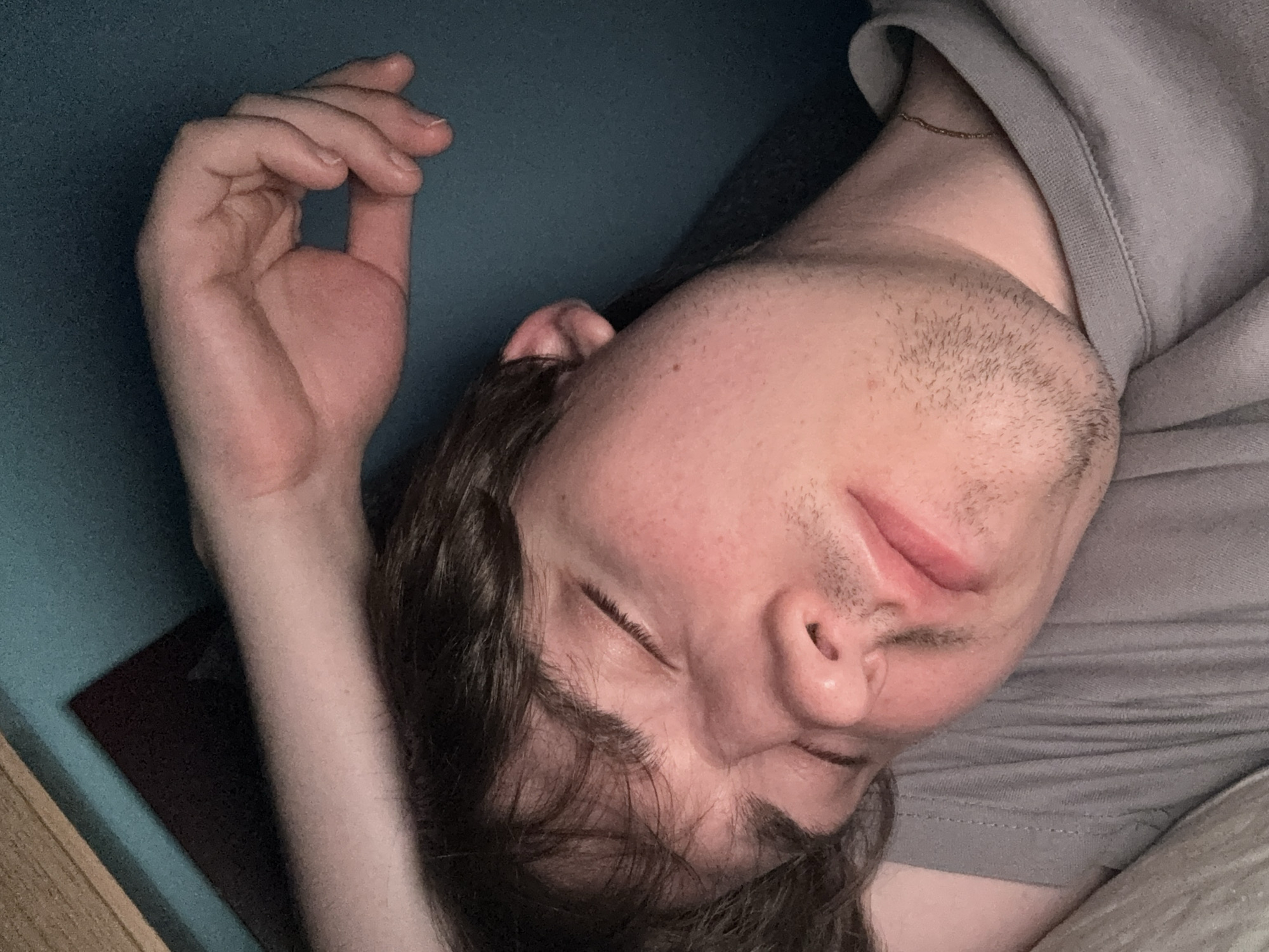 The image shows a close-up, overhead view of a person with their eyes closed, appearing to be asleep. The person has light skin, brown hair that falls across their forehead, and a few days of stubble on their chin and upper lip. They are wearing a grey t-shirt, and a thin gold chain is visible around their neck. Their right arm is raised, with the hand resting near their head. The background consists of a teal-colored wall and a wooden surface at the top left. The lighting is dim, creating shadows on the person's face and highlighting their features. The scene is intimate and peaceful. The image does not provide any information about the location in the city of Dnipro, Ukraine.