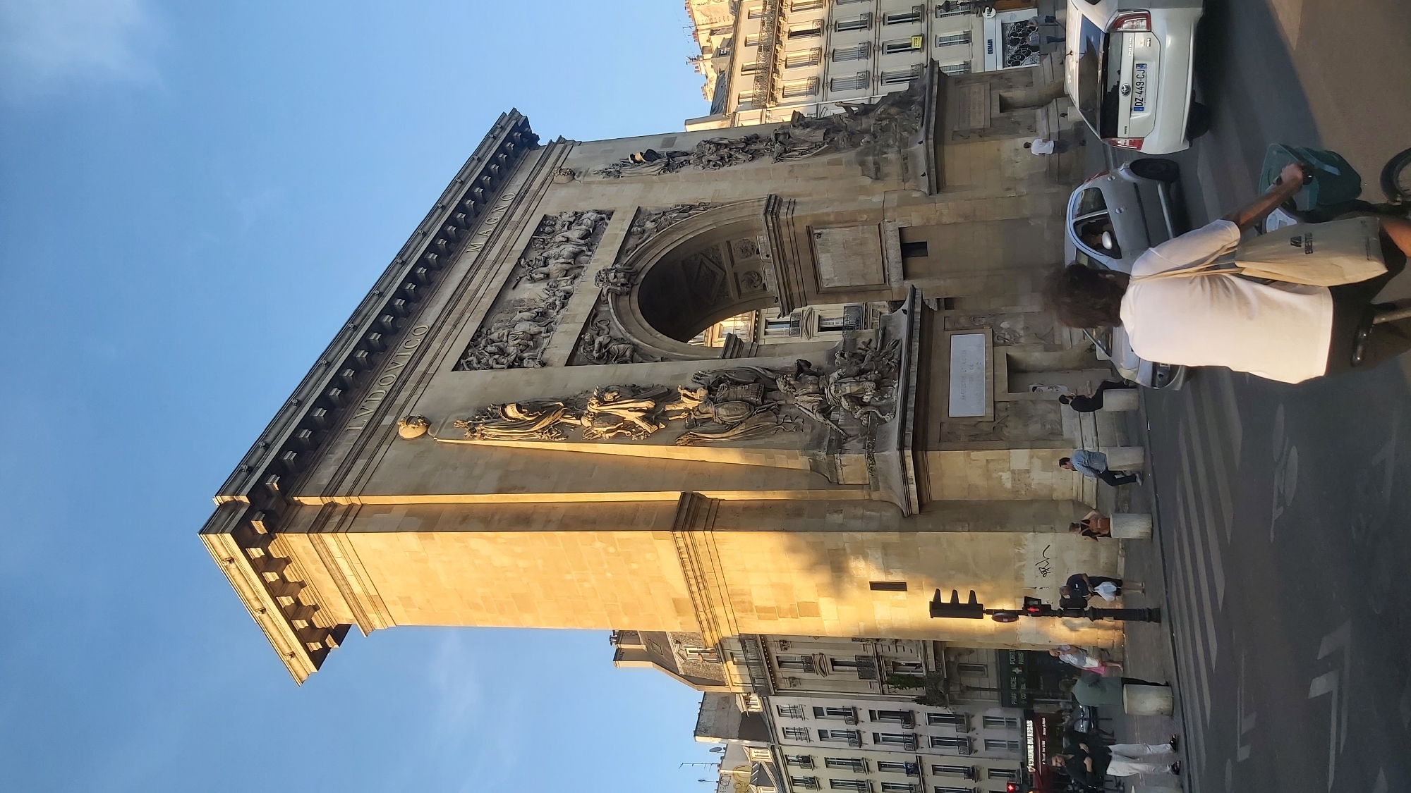The image shows the Arc de Triomphe du Carrousel in Paris, France, bathed in the warm light of the setting sun. The monumental arch, adorned with intricate sculptures and bas-reliefs, stands prominently against a clear blue sky. The inscription "LVDOVICO MAGNO" is visible on the entablature. In the foreground, several people are engaged in various activities. A woman is cycling on the right side of the image, carrying a tote bag. A silver car and a white car are in traffic. Pedestrians are gathered on the sidewalk near the arch, some sitting on bollards. Traffic lights are visible on the left, with one showing red. The architecture of the surrounding buildings, typical of Parisian streetscapes, is also visible. The scene captures a moment of urban life in Paris at dusk.
