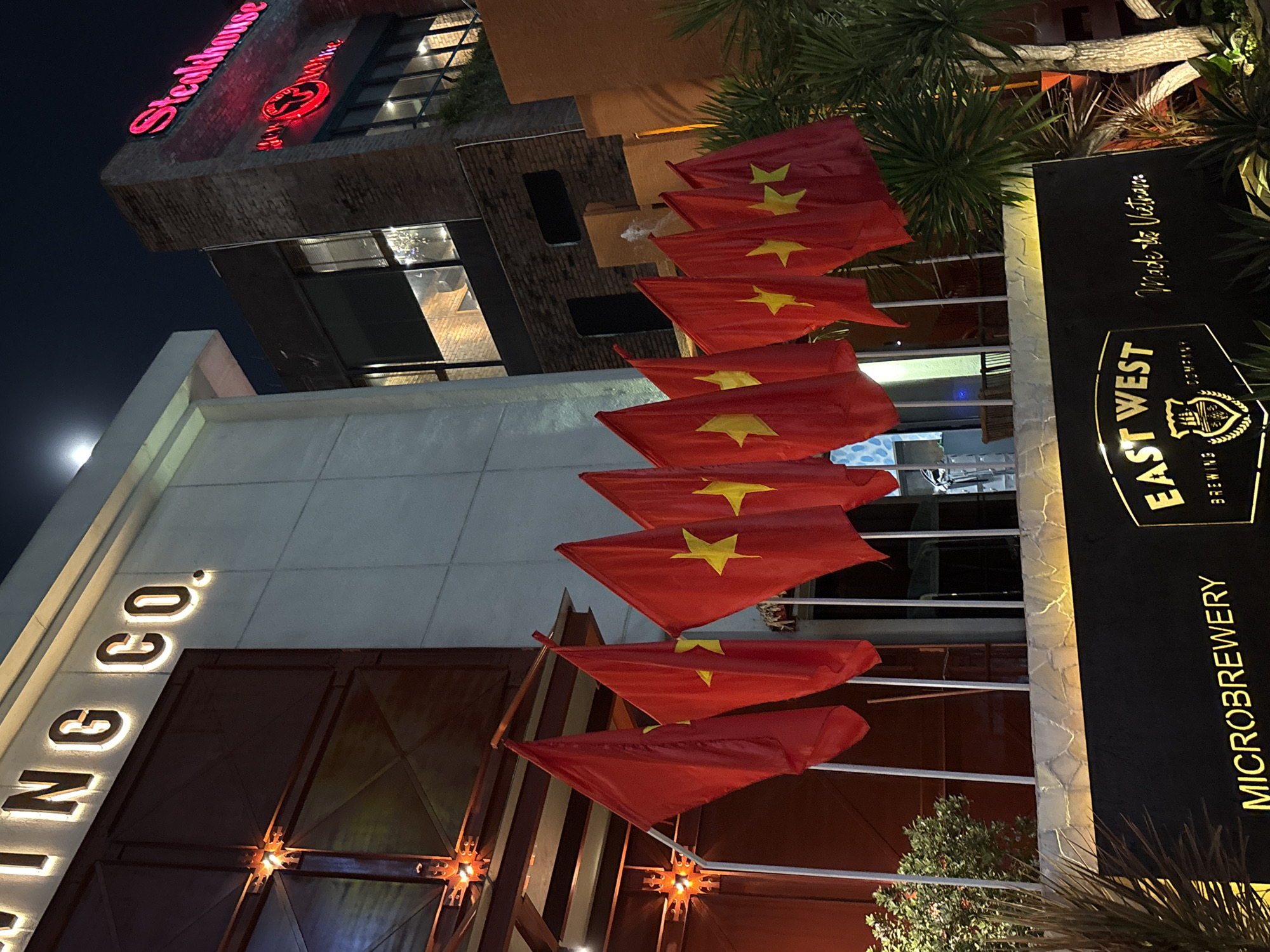 A nighttime shot depicts the exterior of the East West Brewing Co. microbrewery in Đà Nẵng, Vietnam. Multiple Vietnamese flags hang prominently outside. The building's architecture includes a section with illuminated "ING CO." lettering. Further up is a building, marked with "Steakhouse" in red neon lettering. The lighting is dim, with visible neon signs and the moon visible in the sky. The surrounding environment includes trees and foliage.