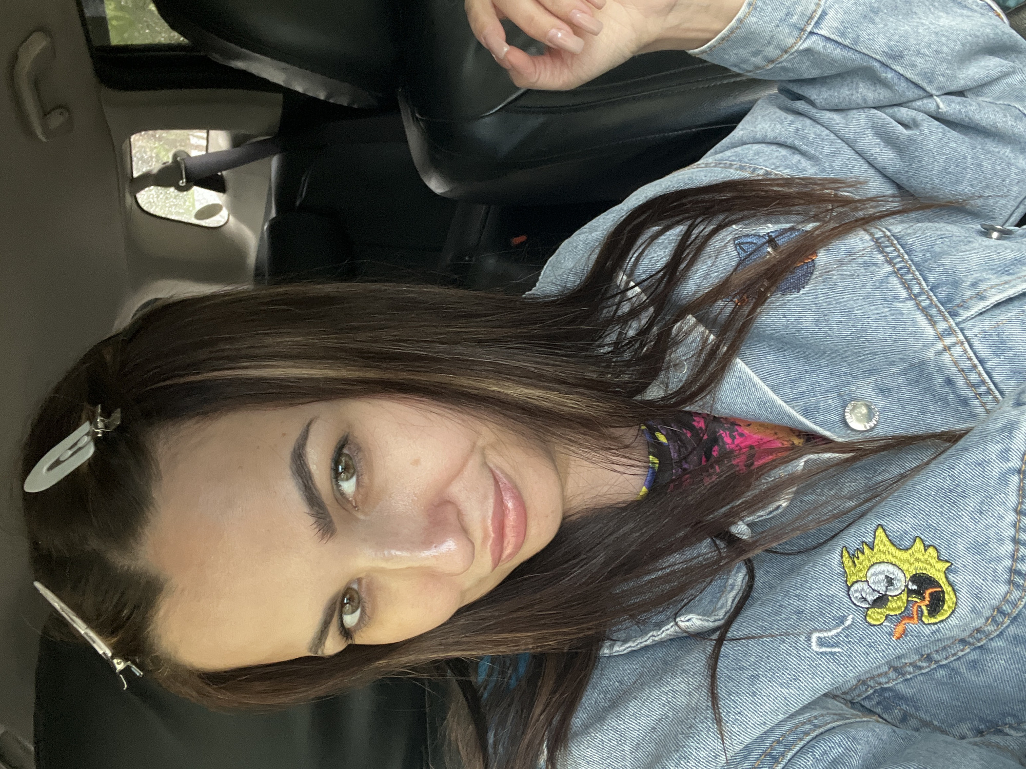 A woman with brown hair is sitting in a car. She is wearing a light blue denim jacket with patches. Her hair is held back with clips near her forehead. The car has black leather seats, and there is a window visible in the background.