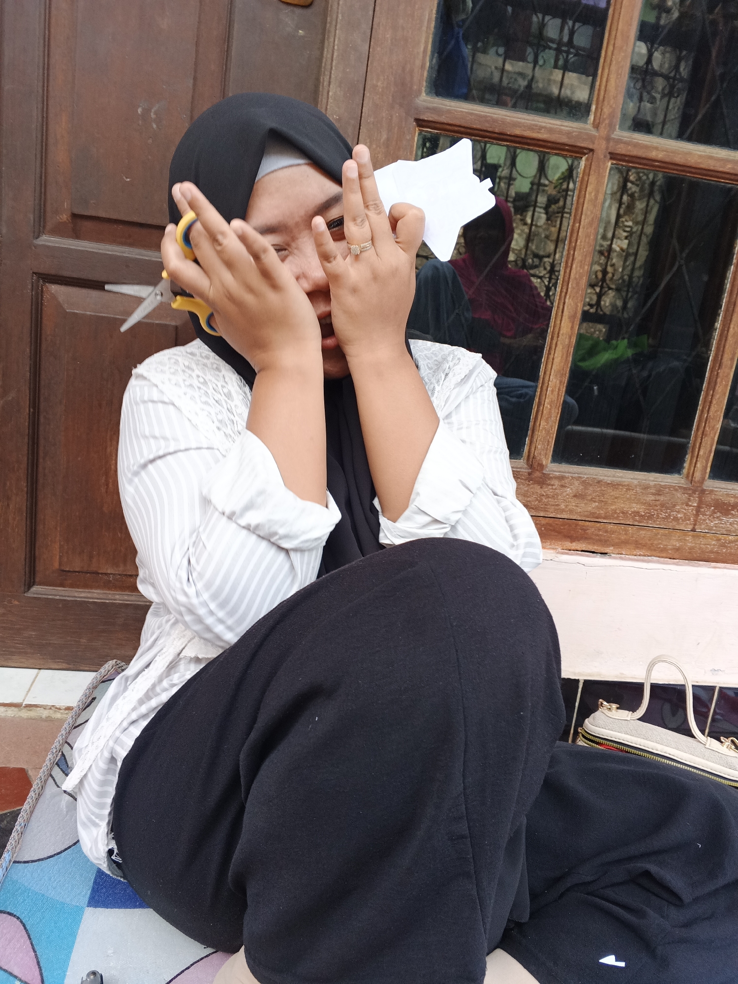 A young woman wearing a black hijab and a white striped shirt is sitting on the floor in front of a wooden door and window. She is holding a pair of yellow-handled scissors in her right hand, and a piece of white paper in her left hand. Her face is partially obscured by her hands, and she appears to be crying or laughing. In the reflection of the window, another person wearing a red hooded jacket can be seen. To the right of the woman, a white handbag with gold accents is visible. The ground beneath her is tiled. The overall setting suggests an outdoor space, possibly a porch or patio, in Warungkiara, Indonesia.