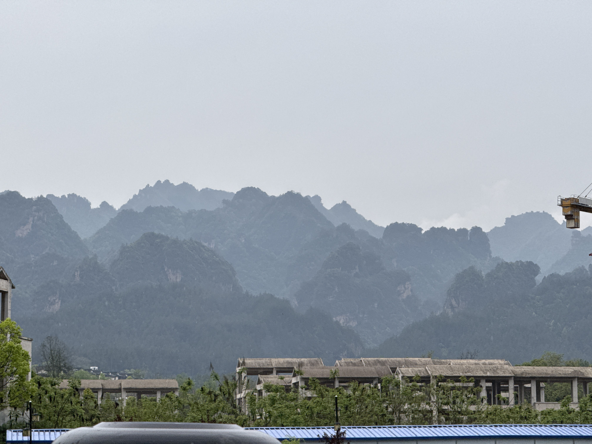 This image captures a panoramic view of a mountainous landscape typical of Wulingyuan, China, under an overcast sky.
The dominant feature is the extensive range of karst mountains that stretch across the mid-ground and background. These mountains appear layered and recede into the distance, rendered in varying shades of grey-blue, suggesting atmospheric haze or mist. Their rugged, craggy peaks and slopes are covered in dense vegetation, giving them a textured appearance. The sky above is a uniform, light grey, indicating an overcast day with diffused light.
In the mid-ground, below the mountains, there are several man-made structures partially obscured by lush green foliage. These buildings appear to be multi-story, possibly residential or industrial, characterized by concrete frames and roofs that seem unfinished or weathered. Some sections have visible windows or open archways.
The foreground contains more elements of human activity and infrastructure. To the far left, a corner of a light-colored building with green trees beside it is visible. Across the bottom of the frame, a bright blue corrugated roof stretches horizontally, likely part of a modern structure. A dark, curved object, possibly the roof or hood of a vehicle, is also present in the immediate foreground, obscuring the view slightly. On the far right, the upper section of a yellow construction crane stands out against the muted mountain backdrop, hinting at ongoing development in the area.
No specific human activity is discernable, and there is no visible text in the image. The overall impression is one of a vast natural landscape coexisting with human development, under quiet, possibly damp, weather conditions.